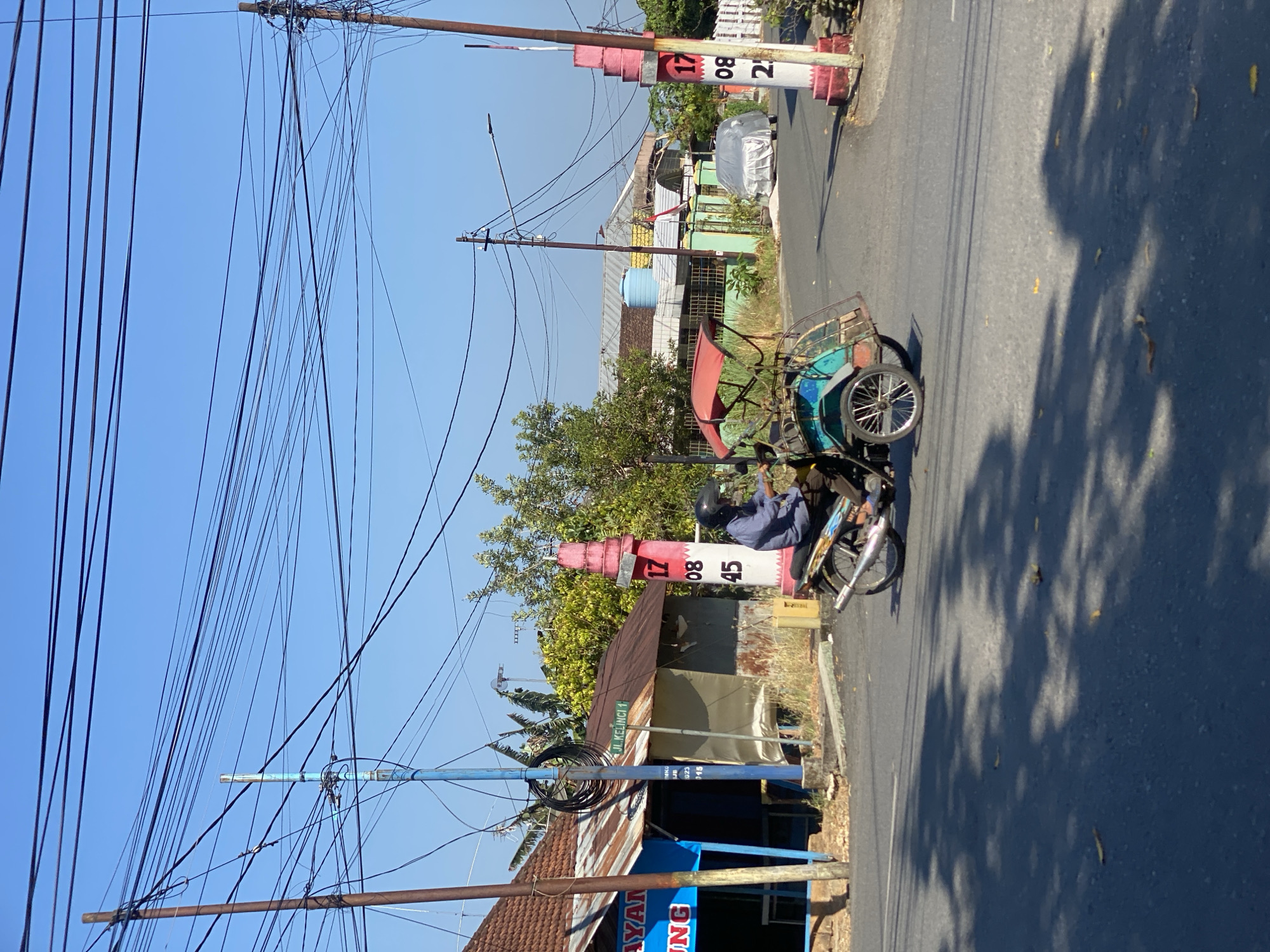 In Banjarbaru, Indonesia, a person rides a motorcycle with a sidecar attached, a common mode of transport known as a "becak motor." The rider wears a blue long-sleeved shirt and a black helmet. The becak motor has a red canopy and is turquoise and red in color. The scene unfolds on a paved road under a clear blue sky, with numerous overhead power lines crisscrossing the frame. To the left of the road, a small building with a rusty corrugated metal roof and a blue awning is visible. A street sign indicates "JL. KELINCI 1". On the right side of the road, a decorative pillar painted with white and red bands stands tall, featuring numbers "17", "08", and "45". In the background, more buildings with tiled roofs and trees can be seen, along with a car covered in a white tarp. The sun casts shadows from trees onto the road, indicating it is daytime.