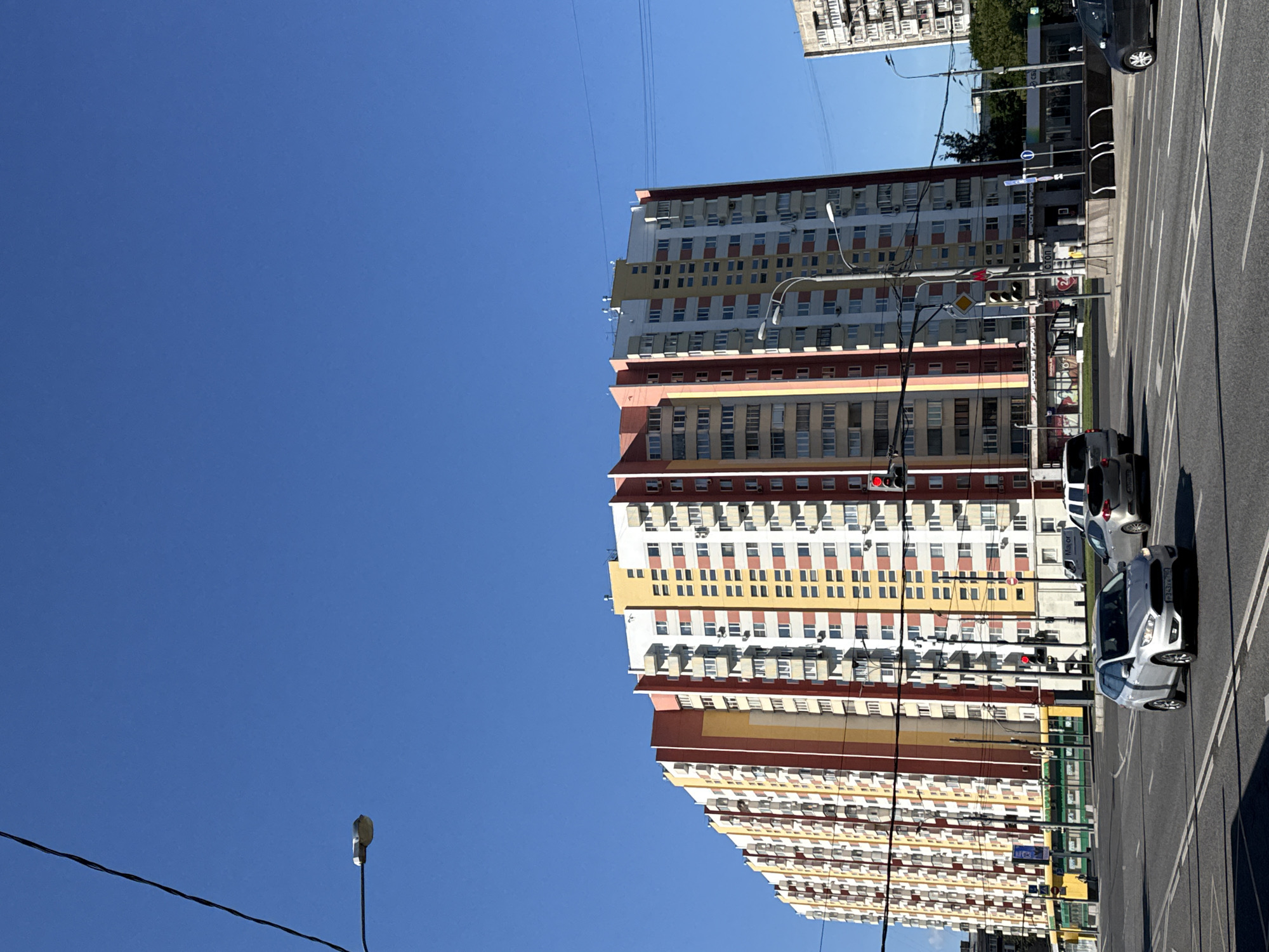 The image shows a wide, multi-lane road in Moscow, Russia, under a clear blue sky. A long, colorful residential building dominates the background, with sections painted in red, yellow, and white. The building has numerous balconies and windows. In the foreground, several cars are driving on the road, including a silver Ford Focus and a white SUV. Traffic lights are visible, one of which is red, indicating a stop. There are also streetlights and power lines crossing the scene. In the mid-ground, a yellow tram or bus is visible to the left, and to the right, there is a building with some shops or kiosks. The overall scene depicts an urban environment on a sunny day.