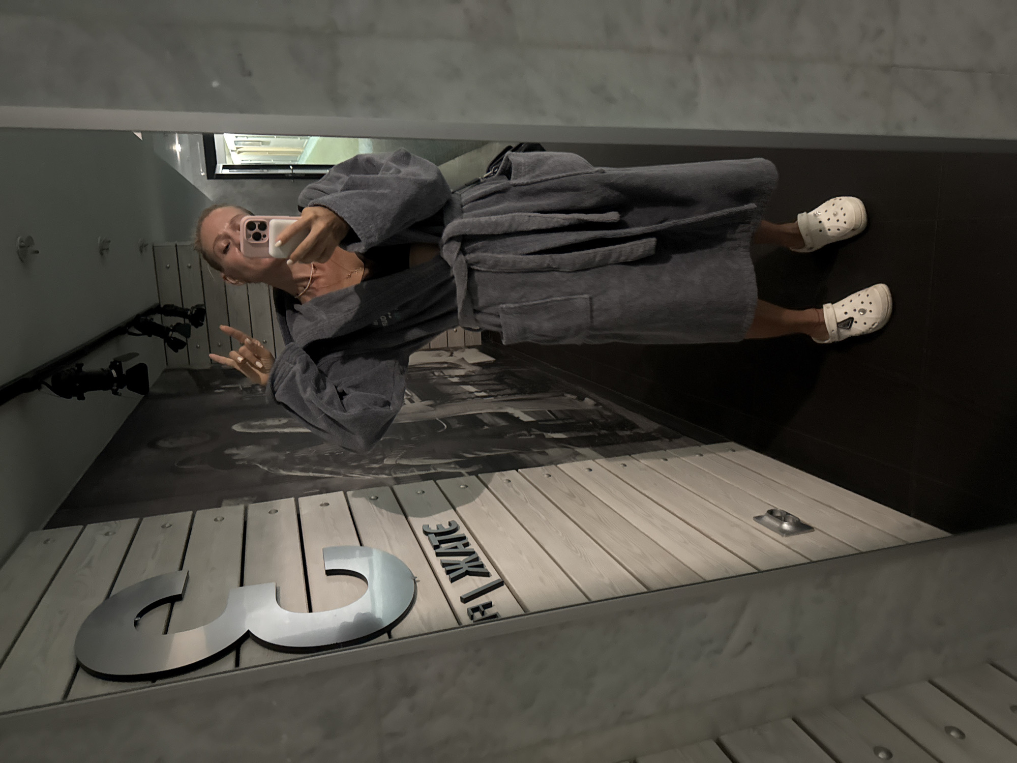 In an indoor setting, possibly a spa or hotel with a modern, minimalist aesthetic, a woman is taking a selfie in a mirror. She is wearing a gray bathrobe and white Crocs, accessorized with a thin necklace and a black bra visible beneath her robe. The background features a large, metallic letter "E" mounted on a light-colored, paneled wall, and a black-and-white mural. The flooring is dark. The overall impression is a casual, relaxed moment captured in a contemporary urban environment. The city of Yalta, Ukraine, is mentioned as the location.