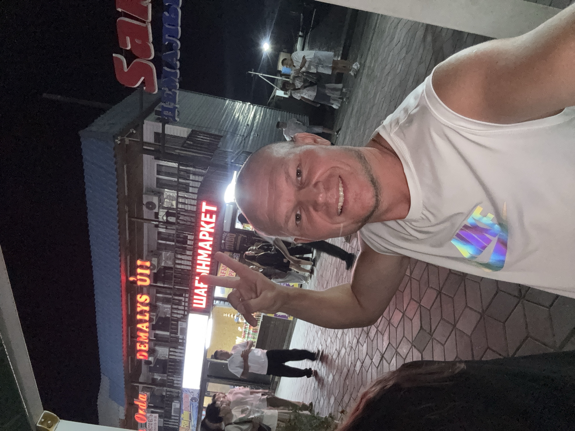 A mid-shot, selfie-style photograph captures a bald, Caucasian man with a shaved head and a light stubble, wearing a white sleeveless t-shirt with a holographic Nike logo on the chest. He is in the foreground, smiling and extending his left arm to make a peace sign with his fingers. The background depicts a nighttime scene with a paved pedestrian walkway. Behind him, a building with illuminated signs is visible. One sign reads "DEMALYS ÚII" in large red letters, and another smaller sign below it says "ШАГІНМАРКЕТ" in red Cyrillic characters. Further to the right, a large red sign with white lettering, partially visible, appears to say "San." Several individuals are present in the background, walking and standing. The environment suggests a commercial or recreational area, possibly a marketplace or entertainment district. The overall impression is a casual, outdoor social gathering at night. The presence of specific signage, such as "DEMALYS ÚII" and "ШАГІНМАРКЕТ", combined with the architectural style and the context of the selfie, are characteristic of locations within or around Zharbulak, Kazakhstan, a city known for its bustling night markets and commercial centers.
