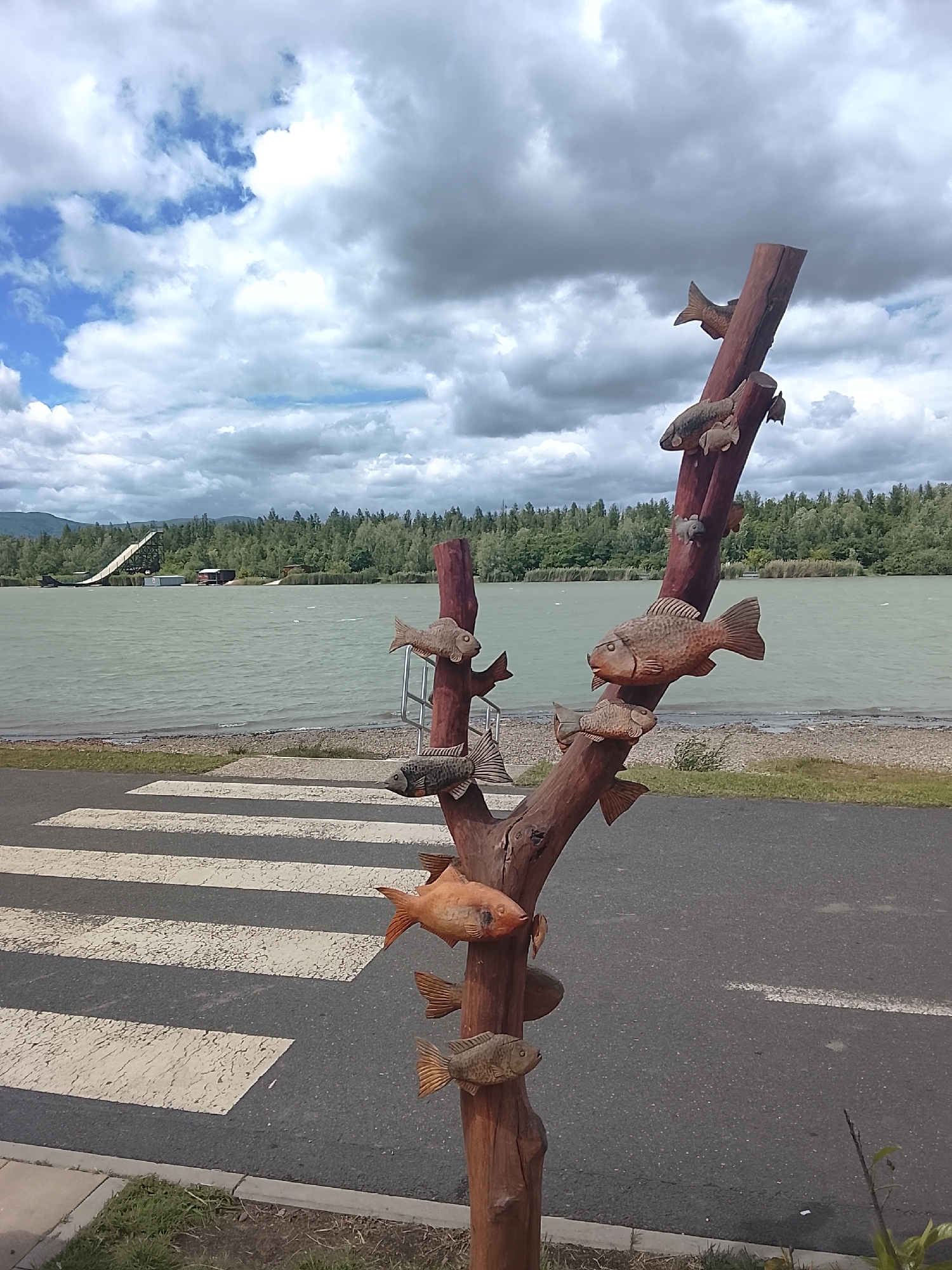 A wooden sculpture of fish is mounted on a wooden pole in front of a lake. The sculpture is located on a paved area next to a crosswalk. The lake is calm, and the sky is cloudy. In the background, there are trees and hills. There is a large ramp structure on the left side of the image, extending into the lake. The overall scene suggests an outdoor recreational area near a body of water.