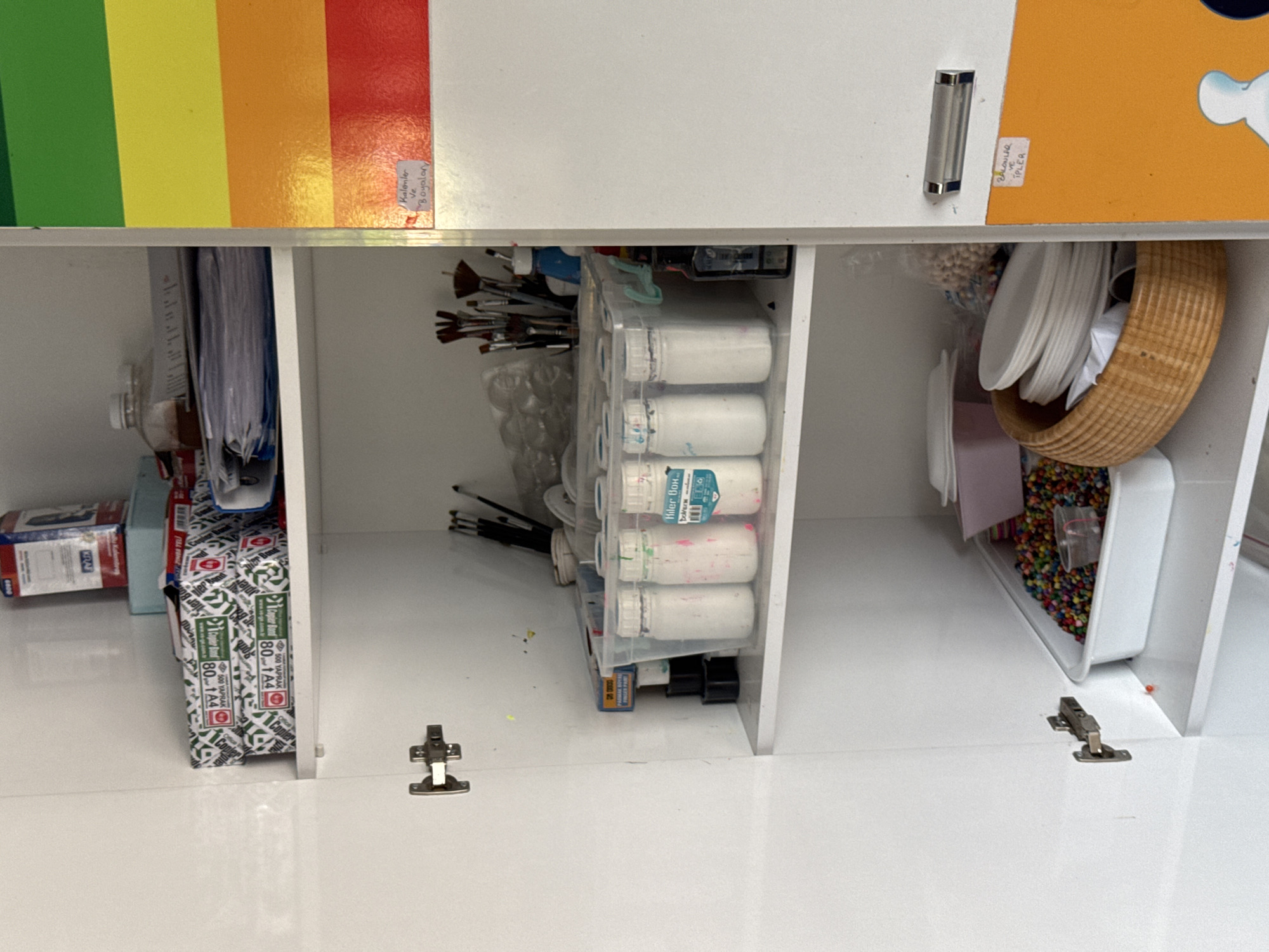 The image shows the interior of a white cabinet, likely in a classroom or art studio, located in Aksu, Turkey. The cabinet is filled with various art supplies.
On the top shelf, there are stacks of paper, a box of what appears to be art supplies, and a document folder.
The middle shelf contains a clear plastic bin filled with bottles of paint. To the right of this bin, there is a collection of paintbrushes in a container.
The bottom shelf holds a white tray filled with colorful beads, a basket with stacked white plates, and some plastic containers.
The right side of the cabinet door displays a colorful chart with horizontal bands of green, yellow, orange, and red. A handwritten note in Turkish, which translates to "Pencils and Paints," is attached to the red band. Another handwritten note in Turkish, translating to "Drawing and Shapes," is visible on the orange band. The cabinet has silver handles on its doors.