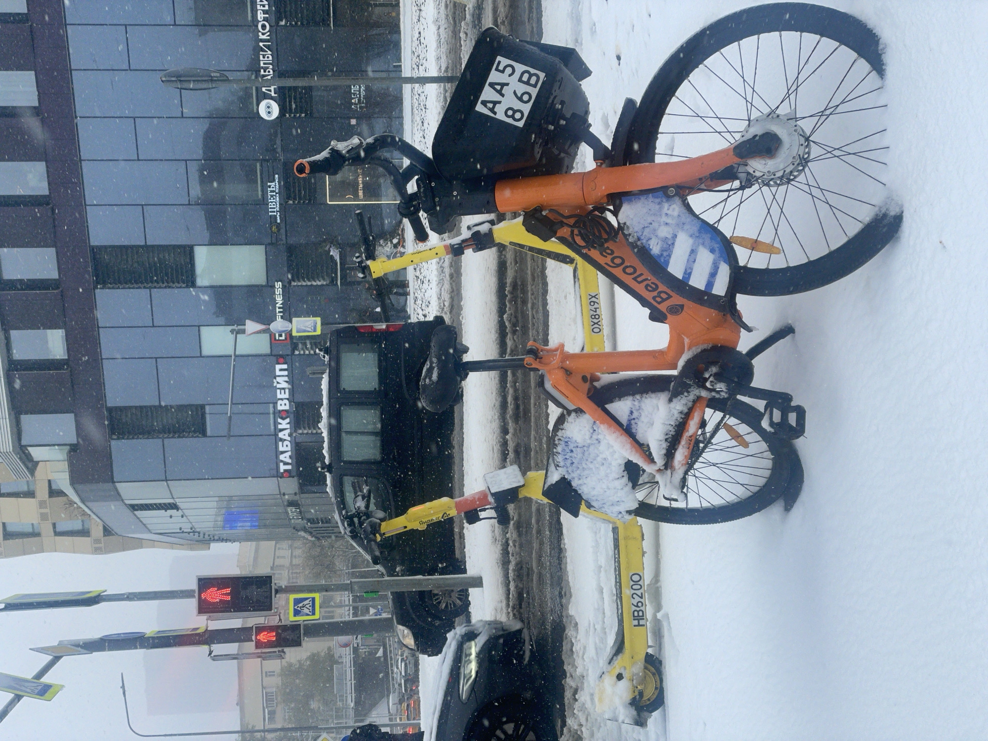 The photograph captures a snow-covered scene in what appears to be a city environment, identified through signage as Khimki, Russia. Two rental bikes are parked on the sidewalk, partially buried in the snow. The first bike, in the foreground, has a registration number "HB6200" on its yellow frame. Above it, another rental bike, with an orange frame, is partially visible with its license plate labeled "AA5 86B". A black container is attached to the front of the orange bike.
In the background, there is a black SUV, with a dark-tinted passenger section. Various street signs are visible, including one with the word "FITNESS," another with the Russian word for "flowers" ("ЦВЕТЫ"), and a sign for "ДАБЛБИ КОФЕЙ" (Doubleby Coffee). Also visible is the phrase "ТАБАК ВЕЙП". Traffic lights display a red pedestrian signal, indicating pedestrians should not cross. Snow is falling, obscuring visibility.