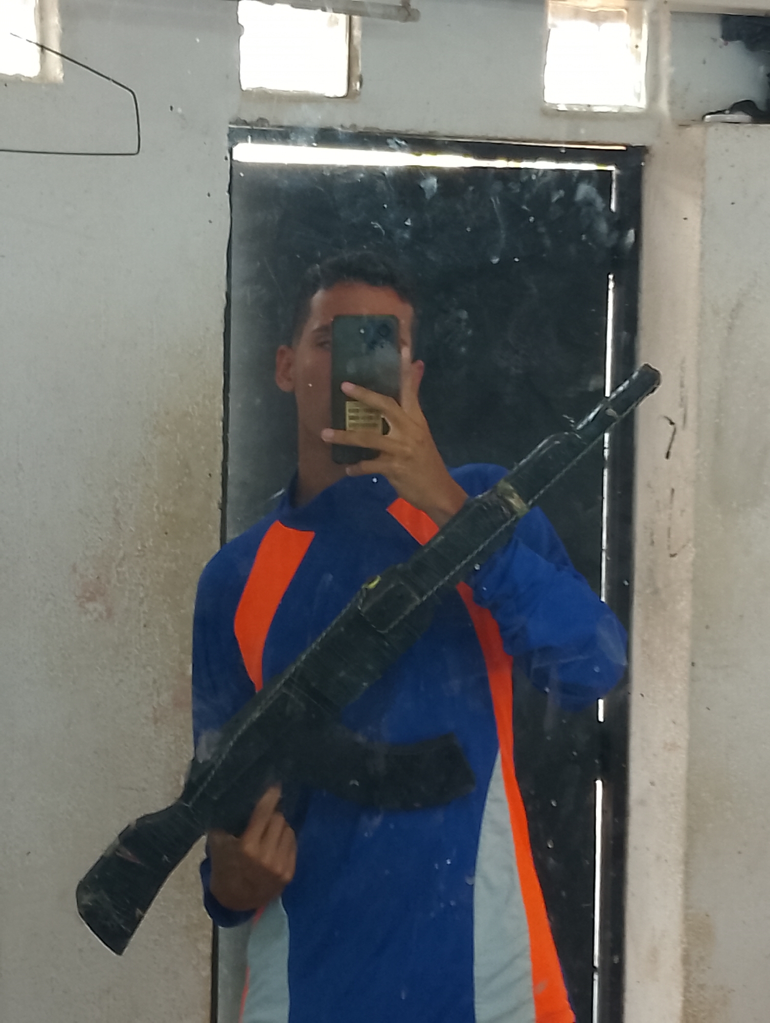 A person is holding a gun in front of a dirty mirror. The person is wearing a blue, orange, and gray long-sleeved shirt and is holding a cell phone to take a selfie. The mirror is in front of a door. The location is Porlamar, Venezuela.