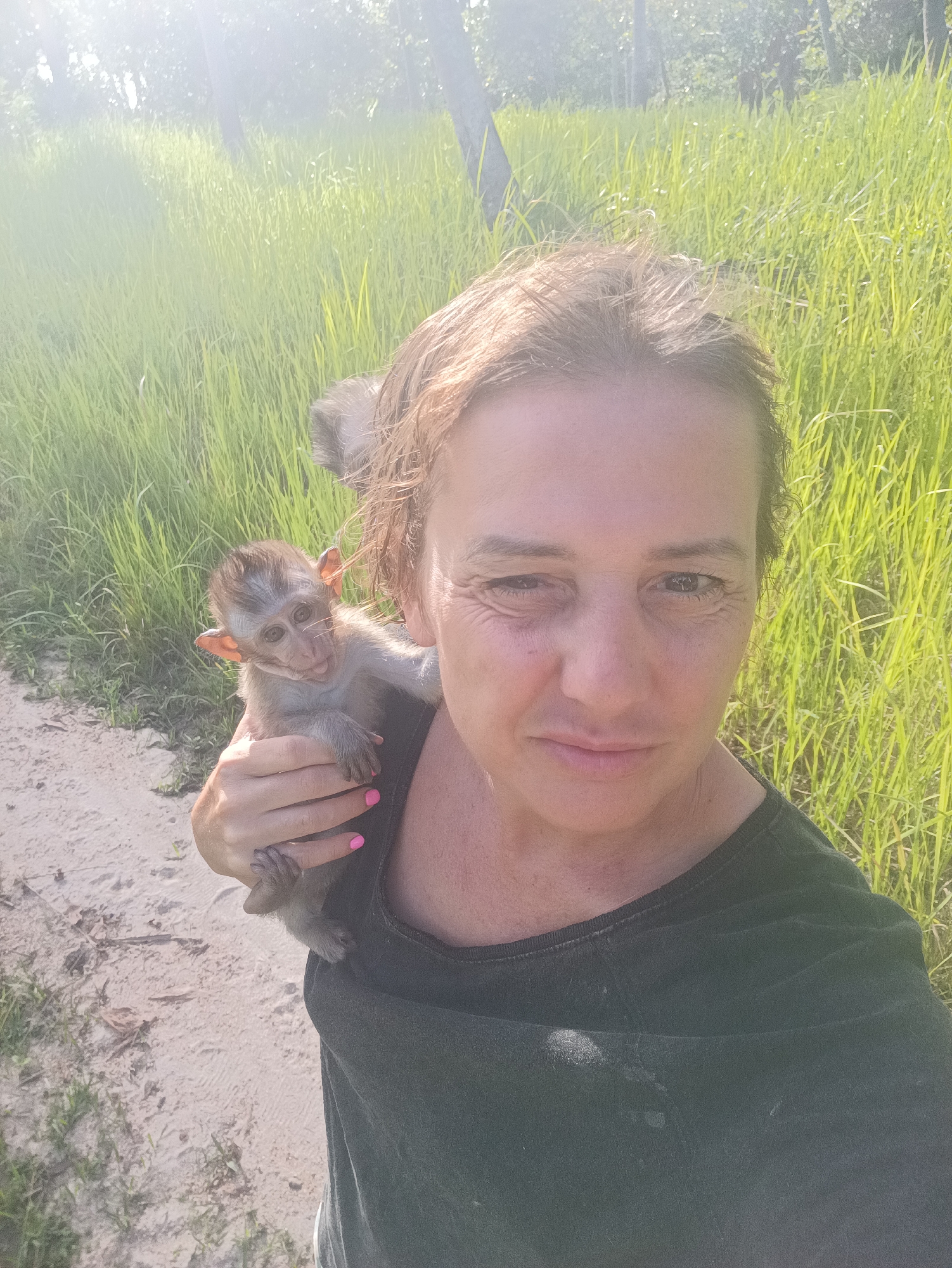 A woman is holding a baby monkey on her shoulder outdoors. The woman has fair skin, light brown hair, and wears a dark green t-shirt. Her fingernails are painted bright pink. The monkey is small and gray, with large ears and light eyes. It has a small tail and grips the woman's shoulder with its hands and feet.
In the background is a path with light-colored sand, dense tall green grass, and trees. The lighting suggests it is daytime. The location is Baan Thong Nang, Thailand.