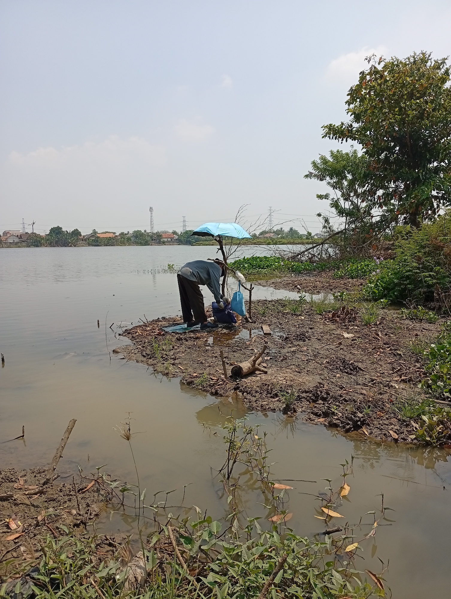 A person wearing a gray long-sleeved shirt and dark pants is standing by the edge of a muddy lake in Karangmukti, Indonesia. The person is hunched over, interacting with equipment placed on the ground. A small, light blue umbrella is propped up behind the person, offering shade. The surrounding environment is natural, with the lake taking up a significant portion of the frame. The shoreline is muddy and covered with dried leaves and sparse vegetation. In the background, across the lake, there are houses and trees, as well as tall power line towers, indicating proximity to a populated area. The sky is clear and pale, with a few faint clouds. The water is calm and reflects the muted colors of the sky and surroundings. There are some aquatic plants growing in the shallower parts of the lake near the shore.