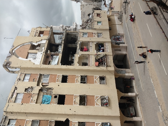 This image depicts a street scene in Khan Yunis, Palestinian Territory, featuring a severely damaged multi-story building under an overcast sky. The building's light-tan facade is riddled with numerous impact marks and bullet holes. Extensive structural damage is evident, particularly on the upper floors and the right side, where large sections of walls and ceilings are missing, exposing interiors that include charred areas and debris. An arched decorative element on the upper right is heavily compromised. Several windows are broken or missing, with some covered by blue tarps or white sheets. Laundry hangs from balconies on the middle floors. On the ground level, a white sedan is parked under an arched recess, and a few children are present. The paved street features a central white line, flanked by dirt verges. A group of approximately five individuals, predominantly adults, are seated or crouching on the dirt patch to the right of the street. One individual in an orange and black jacket stands in the roadway, and another person pushes a cart further down the street on the far right. In the background, additional damaged buildings, piles of rubble, and a partial mosque dome are discernible.