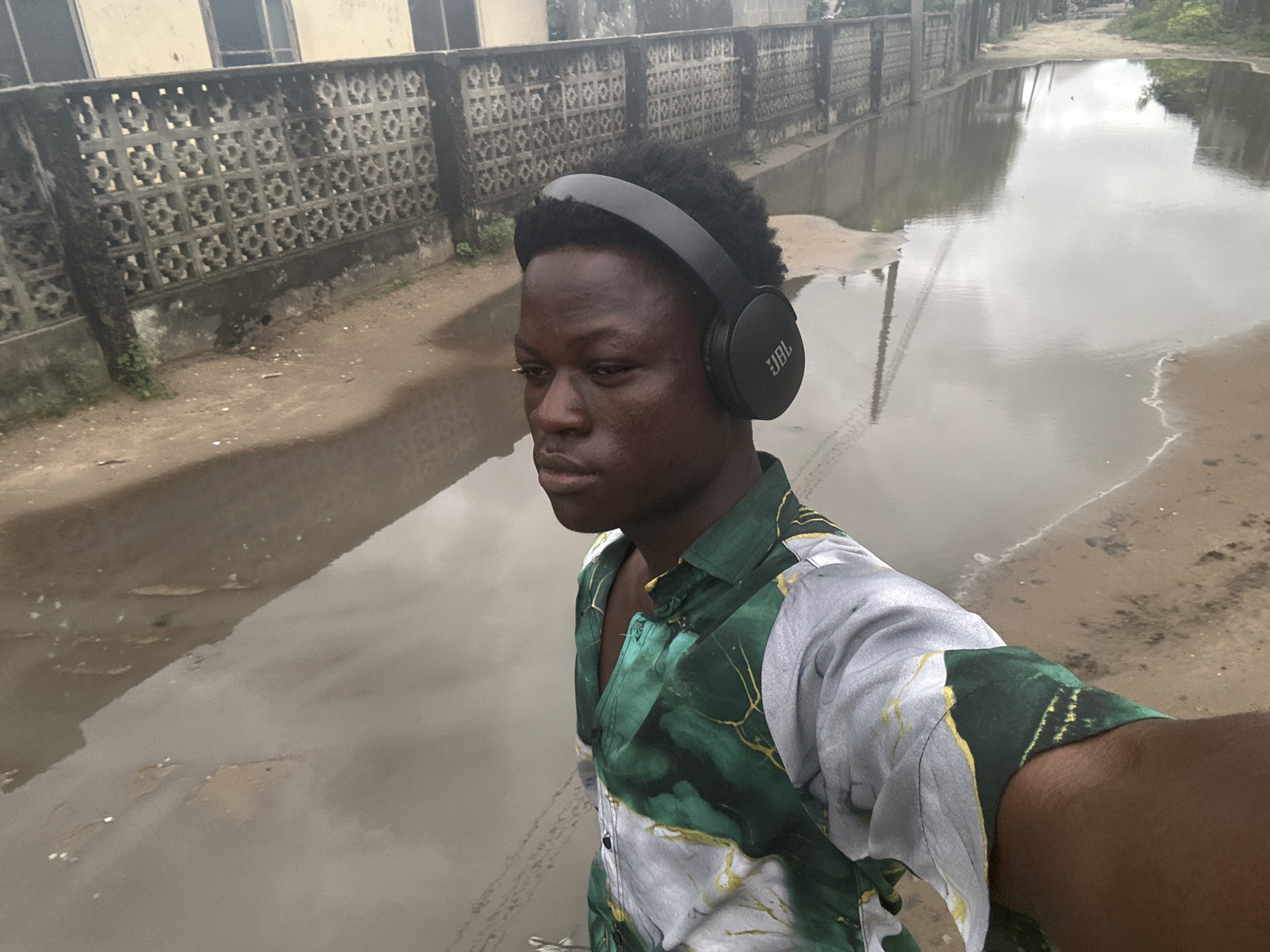 The image features a young Black person wearing black headphones and a green and white patterned shirt. They are positioned in the foreground, facing slightly to the left, with their head turned towards the camera. The scene appears to be outdoors in a wet, possibly post-rain environment, with large puddles on a sandy or dirt road. In the background, there's a textured concrete wall with a repetitive geometric pattern, and beyond that, indistinct buildings. The sky is overcast, suggesting a cloudy day. The reflection of the sky and surrounding elements is visible in the water. The overall mood is somewhat casual, possibly indicating the subject is out for a walk or enjoying music despite the damp conditions. The headphones have the "JBL" logo visible on the right earcup. The location is identified as Ajara, Nigeria.