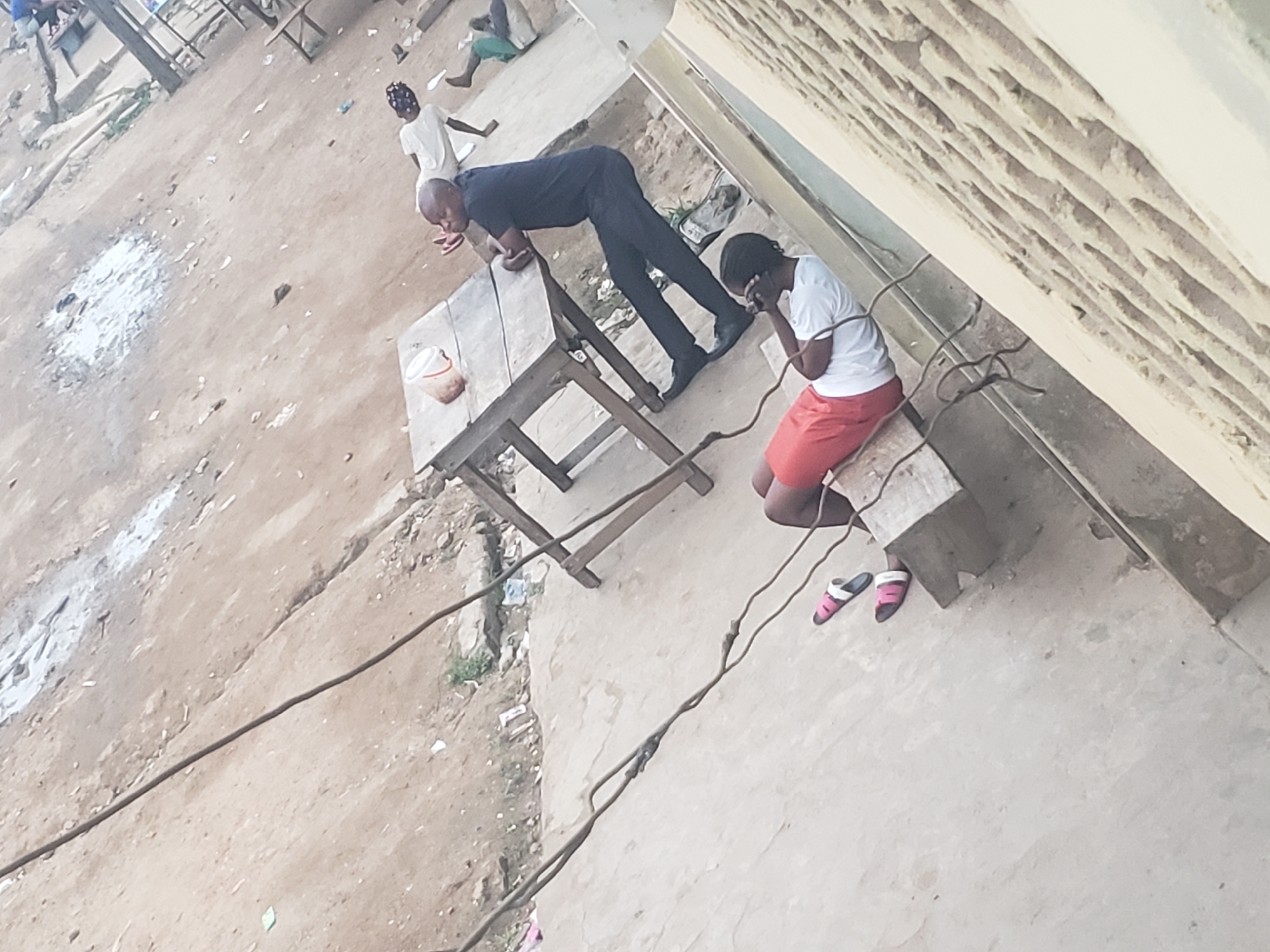 The image depicts a scene in Alalubosa, Nigeria. A man in a dark suit is leaning over a wooden table. Nearby, a woman in a white shirt and orange shorts is kneeling on a concrete slab, with a pair of sandals next to her. An electrical wire runs across the ground. Another person is visible in the background, wearing a white shirt. The surrounding environment appears to be an outdoor area with a dirt ground.