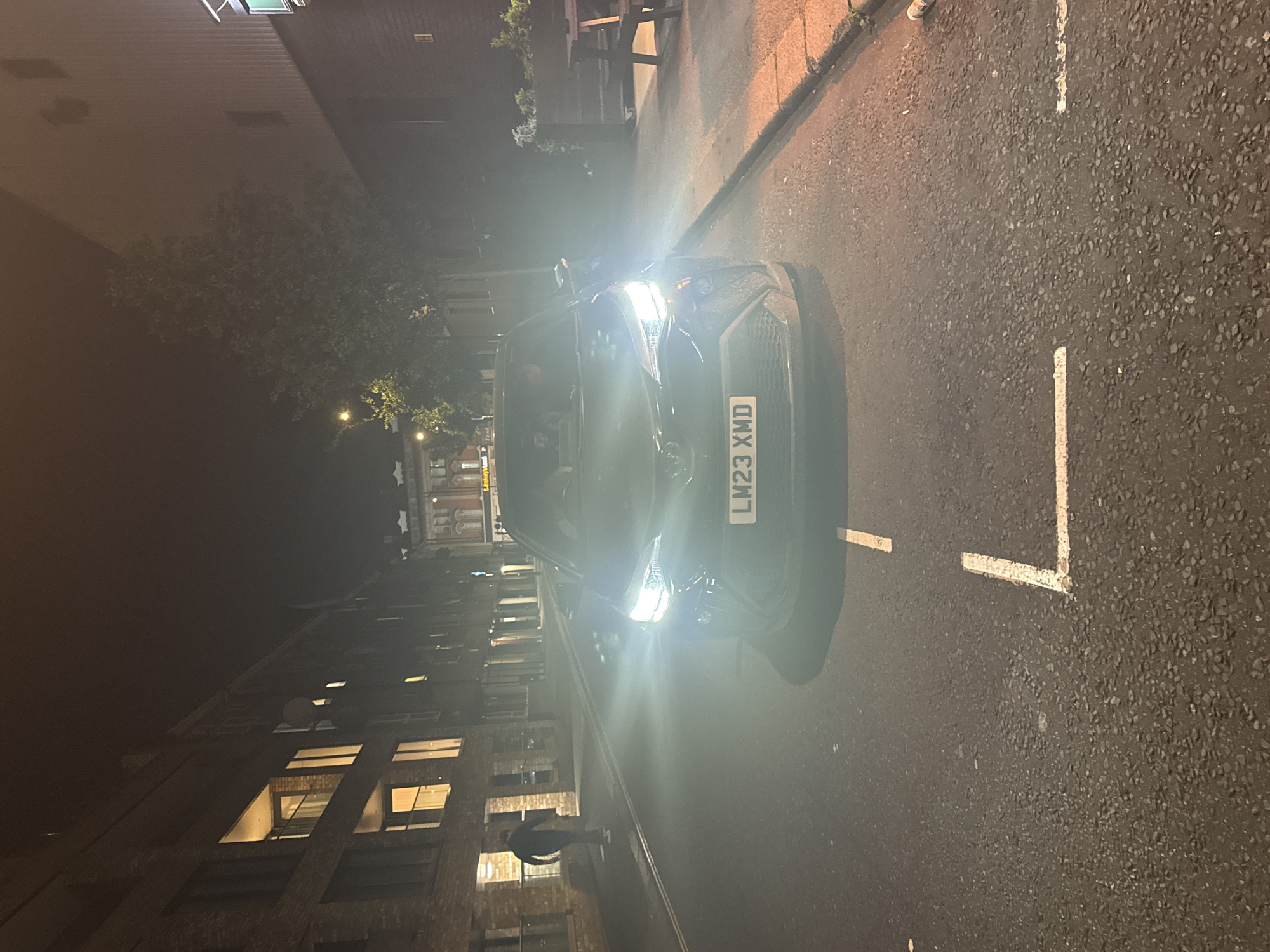 The image depicts a dark urban street scene at night, likely in London, United Kingdom, based on the license plate "LM23 XWD" on the front of a parked black car. The car is positioned on the right side of the frame, with its headlights illuminating the road ahead. The road is marked with white lines indicating parking spaces.
On the left side of the frame, there are buildings with illuminated windows, suggesting residential or commercial properties. A person is walking near the buildings on the left. Above, there is a dim sky, which might be slightly illuminated because of city lights. A tree is seen on the opposite side of the street. The overall impression is of a quiet, dimly lit street in a city at night.