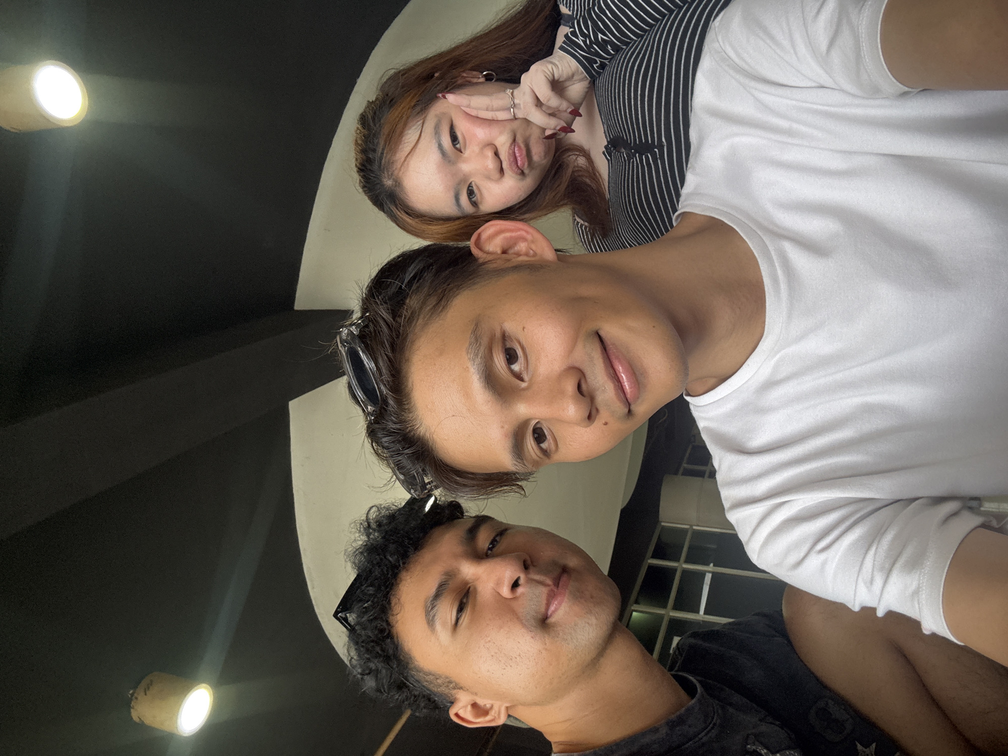 Three people are posing for a selfie indoors, likely in a cafe or restaurant in North Jakarta, Indonesia, given the presence of a pendant lamp and a black ceiling with visible beams, common in such establishments.
In the foreground, a man with curly dark hair and sunglasses perched on his head is on the left, looking directly at the camera with a slight smile. He is wearing a dark t-shirt. Next to him, a man with light brown hair and sunglasses on his head is in the center, also smiling at the camera. He is wearing a white t-shirt. To the right, a woman with long, light brown hair is making a pouty face and holding her hand up to her cheek. She is wearing a black and white striped shirt. The background shows a modern interior with large windows and a dark ceiling with light fixtures.