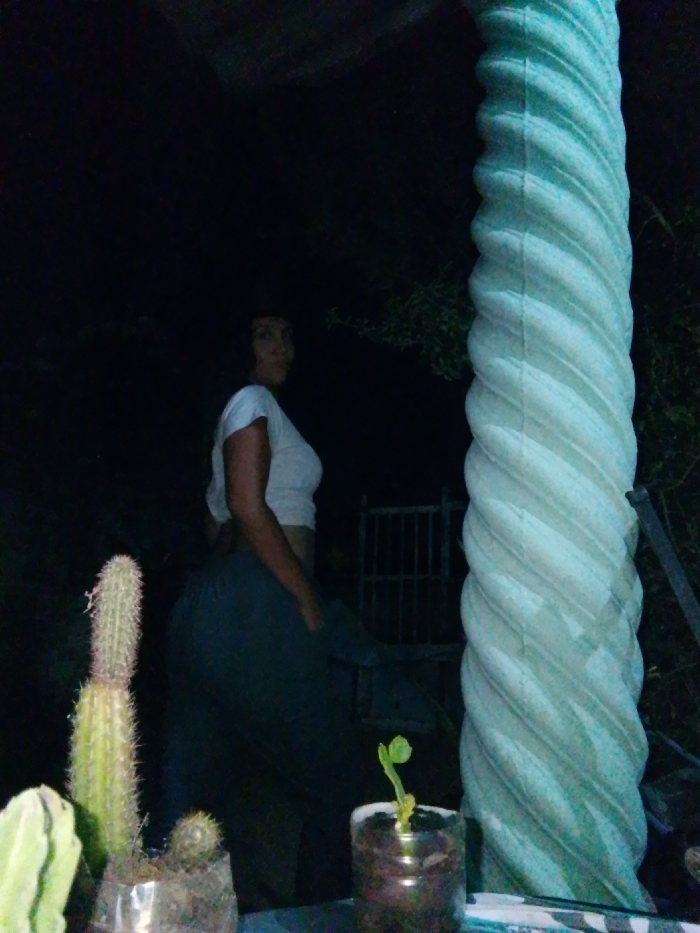 This image captures an outdoor nighttime scene in Juan Griego, Venezuela. An adult female stands in the mid-ground, positioned to the left of a prominent architectural column. She is primarily turned away from the camera, facing right, but her head is rotated to look over her left shoulder towards the viewer. She has dark hair and is wearing a light-colored, short-sleeved crop top and dark, loose-fitting pants. Her right arm is visible with the elbow bent. To the right, a large, light-colored, spiral-fluted column extends vertically through the frame. In the immediate foreground on the left, a tall, spiny cactus and a smaller, round cactus are situated in clear plastic containers. A small plant in a plastic bottle is also visible in the foreground, to the left of the column's base. The background is dark and indistinct, featuring silhouettes of foliage and a metal gate structure between the woman and the column. The scene is dimly illuminated.