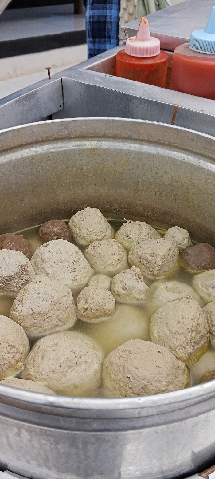 The image shows a large pot of meatball soup, located at coordinates -6.973, 107.52 in Kecamatan Kutawaringin, Indonesia. The pot is made of aluminum and contains approximately 15 meatballs. The soup is a light brown color, and the meatballs are all round and smooth. Two plastic bottles of sauces are visible in the background, one red and one blue. One bottle contains a red sauce and has a pink lid, the other bottle contains red sauce and has a blue lid. There is a rusty nail on the side of the pot. The background of the photo shows the leg of a person wearing a plaid shirt. The scene appears to be outside, possibly a street food stall.