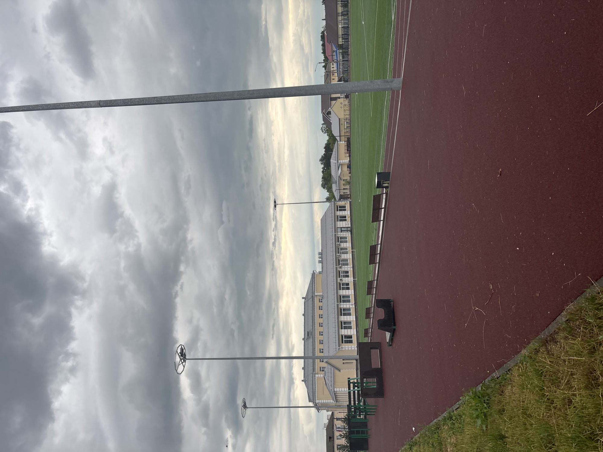 The image depicts an outdoor sports facility on an overcast day in Inskoy, Russia. In the foreground, a red synthetic track runs diagonally from the lower left to the upper right. To the left of the track, a strip of green grass with some weeds is visible. Further into the image, the track transitions to a green synthetic turf field, likely a soccer or sports field, marked with white lines.
Along the edge of the field, several benches are present. To the left of the field, near the track, are some black and green outdoor exercise equipment structures. Two tall, silver lampposts are prominent in the scene, one centrally located and taller than the other to the left. In the background, a long, light yellow building with a grey roof and numerous windows stretches across the landscape. Beyond the building, smaller houses with tiled roofs are visible, and a line of trees can be seen in the distance. The sky is heavily clouded with dark grey formations, suggesting a potential for rain. No people are visible in the image.