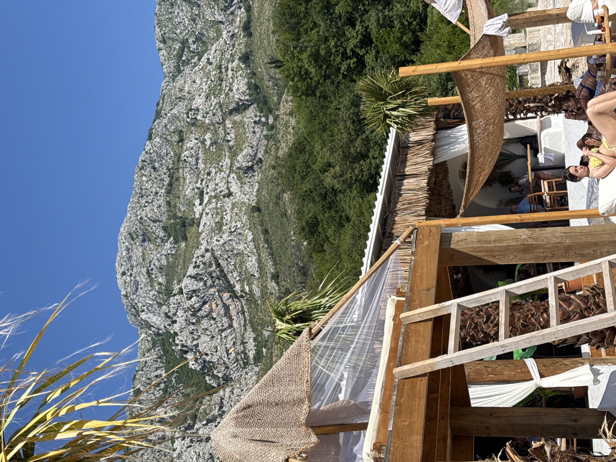 This image depicts a sunny outdoor scene at a coastal resort in Drobnići, Montenegro, with a prominent rocky mountain range in the background.
In the immediate foreground on the left, an out-of-focus, yellow-green palm frond partially obscures the view. Central to the foreground is a rustic, multi-level structure constructed primarily of light-colored wood. Its sloped roof is partially covered by a coarse, tan-colored woven burlap fabric and partially by a transparent plastic sheet. A wooden ladder with four visible rungs leans against a vertical support beam of this structure. Below the structure, white fabric drapes are tied back. Exposed palm tree trunks are incorporated into the architectural design as natural elements.
To the right of this structure, another white building with a thatched roof is visible, featuring white draped curtains. Inside this shaded area, several individuals are seated at tables, their forms partially obscured.
In the bottom center-right, two adult females are depicted lounging on white chaises on what appears to be a sandy or pebbled beach area. The woman on the left, wearing a yellow two-piece swimsuit, is reclining with her legs extended, looking towards the right. The woman to her right, wearing a blue swimsuit, is also reclining, partially obscured by the first woman. A small, low, rustic wooden table, holding a bottle and other indistinct items, is positioned between them.
The midground features a dense band of lush, vibrant green trees and shrubs, creating a natural border between the resort structures and the mountainous terrain.
Dominating the background is a rugged, light grey and white rocky mountain range, characterized by exposed rock faces and sparse patches of green vegetation. Above the mountains, a clear, brilliant blue sky extends across the top of the image, devoid of clouds. The scene is illuminated by bright, direct sunlight, indicating clear weather conditions during daytime.