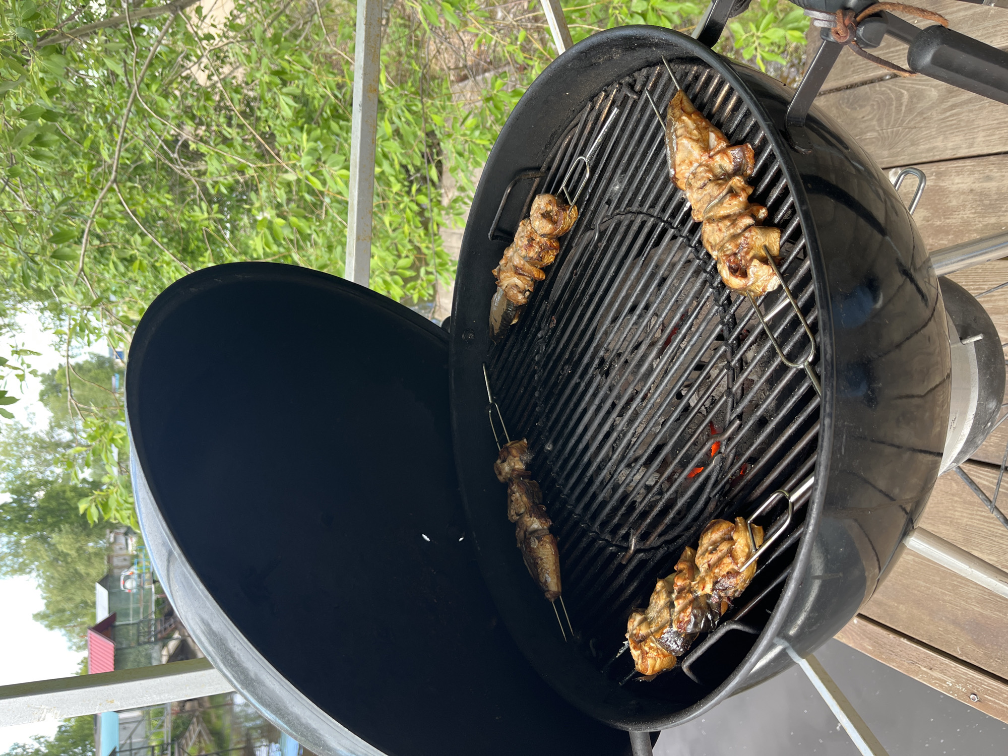 A close-up, eye-level shot shows four skewers of grilled fish on a black kettle grill, placed on a wooden deck. The fish skewers are arranged in a diamond pattern on the grill grates. Some glowing embers are visible beneath the grates, indicating the grill is hot. The background features lush green trees and foliage, with a body of water and small buildings visible in the distance. The scene appears to be outdoors, possibly in a suburban or rural setting, with a focus on the act of grilling. The image does not contain any identifiable individuals.