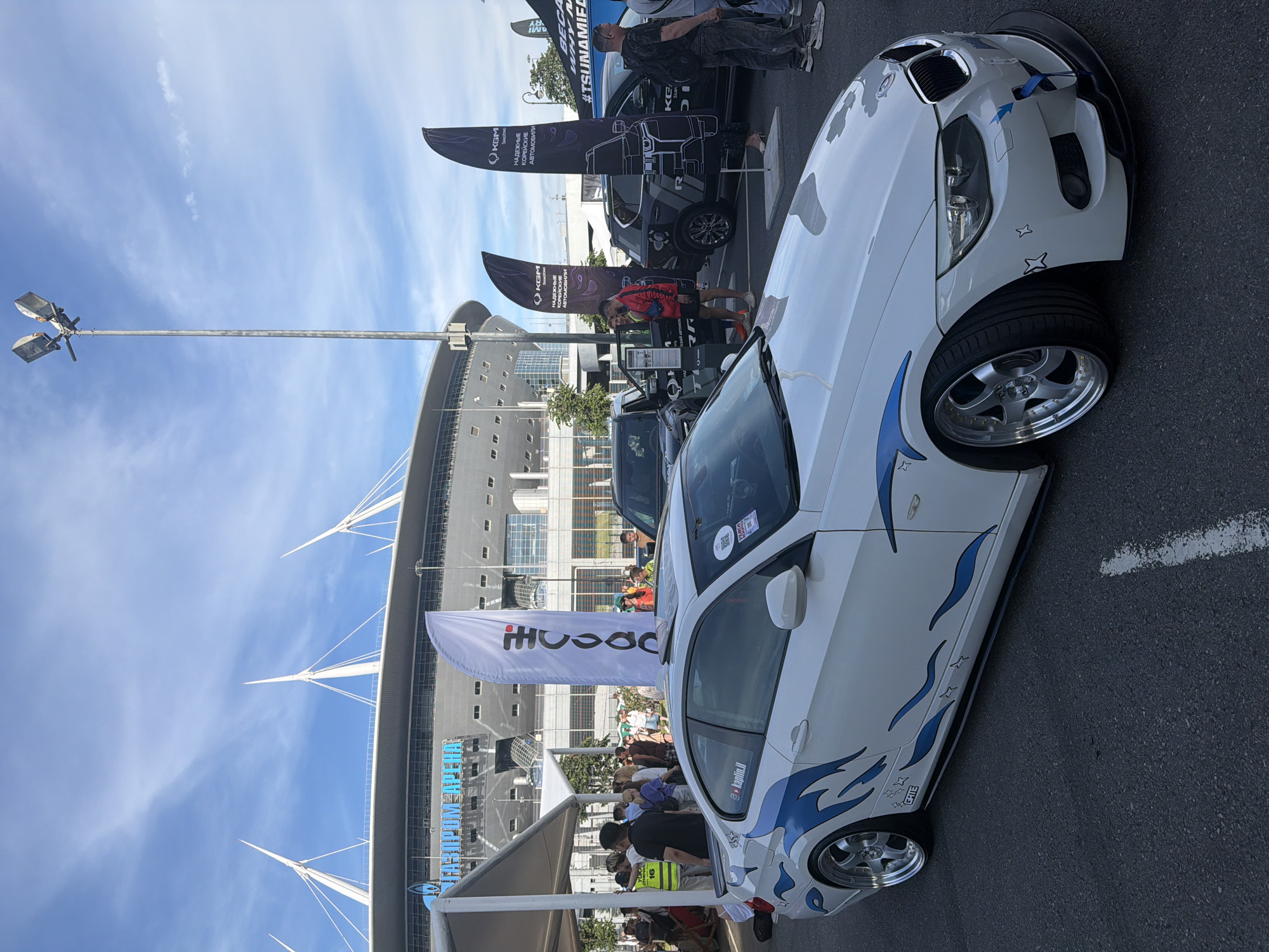 In the city of Unknown, Unknown, a white modified BMW coupe is parked on asphalt in front of the Gazprom Arena stadium. The car features blue flame decals and custom wheels. Several people are gathered around the car and in the background, near several large flags. One flag is white with black text and a red dot. Other flags are dark purple with white text and graphics, promoting SsangYong vehicles. The sky is partly cloudy and blue.