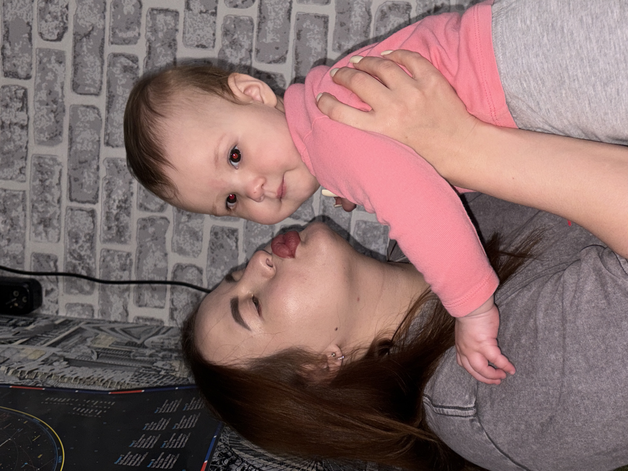 A woman and a baby are lying down. The woman is wearing a gray shirt. The baby is wearing a pink long-sleeved shirt and gray pants. The woman's face is close to the baby's. The background is a gray brick wall. A desk with a black device, and a pad with a design on it, are partially visible on the lower-left side. The image appears to be taken indoors. There is no indication of this picture being taken in Novopokrovskaya, Russia.