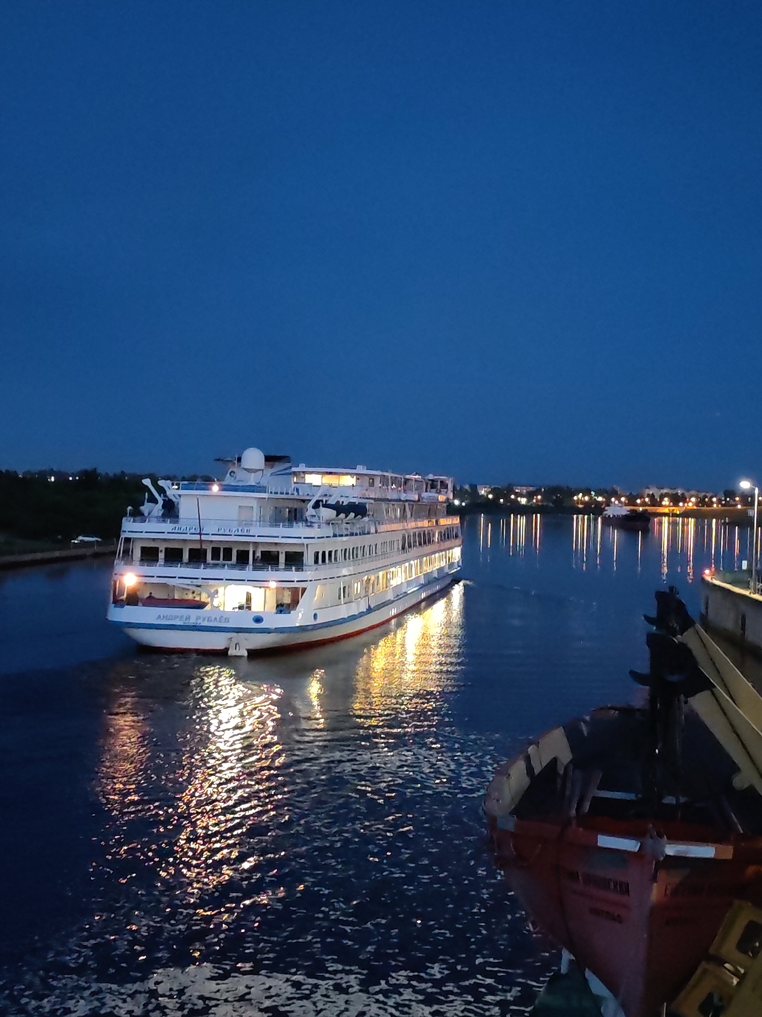 A large white cruise ship, named "ANDREY RUBLEV" in Russian, travels down a calm body of water at dusk. The ship has multiple decks with illuminated windows and is casting reflections on the water. In the foreground, a smaller red boat with dark railings is partially visible, also in the water. The sky is a deep blue, transitioning to darker hues as night approaches. In the background, the lights of a distant city or town are visible along the shoreline, along with trees. A street lamp is also visible on the right side of the frame. The scene appears to be a waterway in or near the city of Sheksna, Russia, possibly a canal or river given the urban and natural elements.