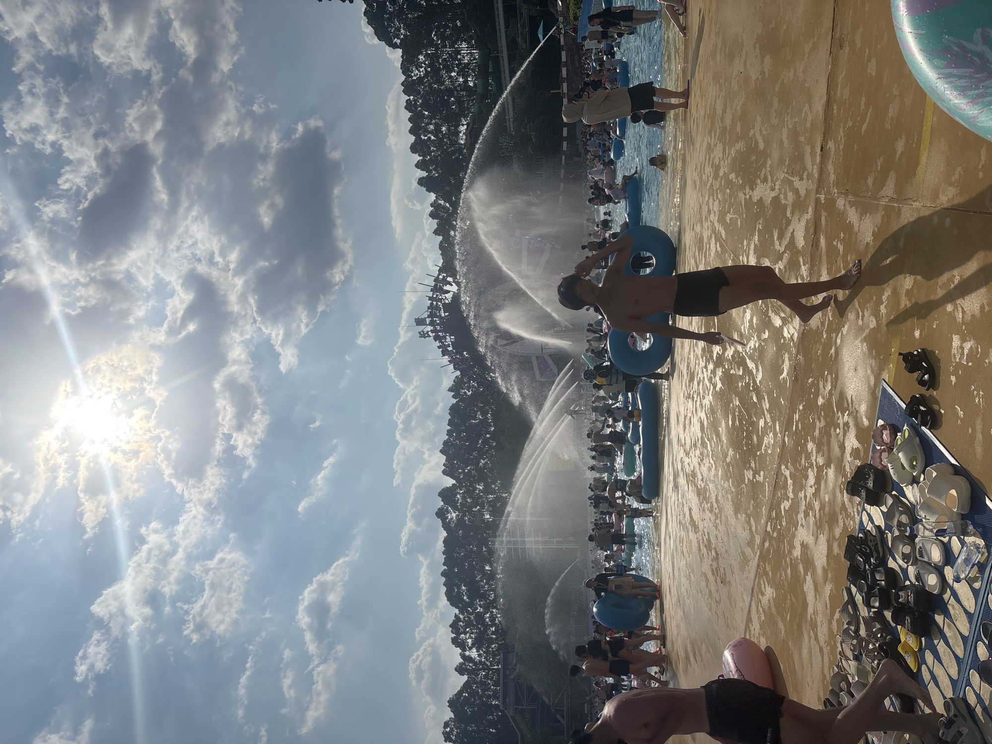 The image depicts a crowded water park in Panyu District, China, on a sunny day. The sky is bright with scattered clouds, and the sun is partially obscured by them, casting lens flares. In the foreground, a sandy area is wet and reflects the sunlight. A large group of people are enjoying various attractions in the water. Several individuals are standing on the wet ground, with one person in the foreground walking towards the right, wearing black shorts and bare-chested. Another person, wearing a light brown hooded sweatshirt and black shorts, stands in the water to the right of the walking individual. In the background, large water fountains are spraying water high into the air, creating a mist. Numerous inflatable rings of various sizes are floating in the water, and many people are seen sitting or floating on them. Trees form a dense line along the horizon behind the water park structures. A collection of footwear, including sandals and flip-flops, is neatly arranged on a blue mat in the lower left portion of the image.