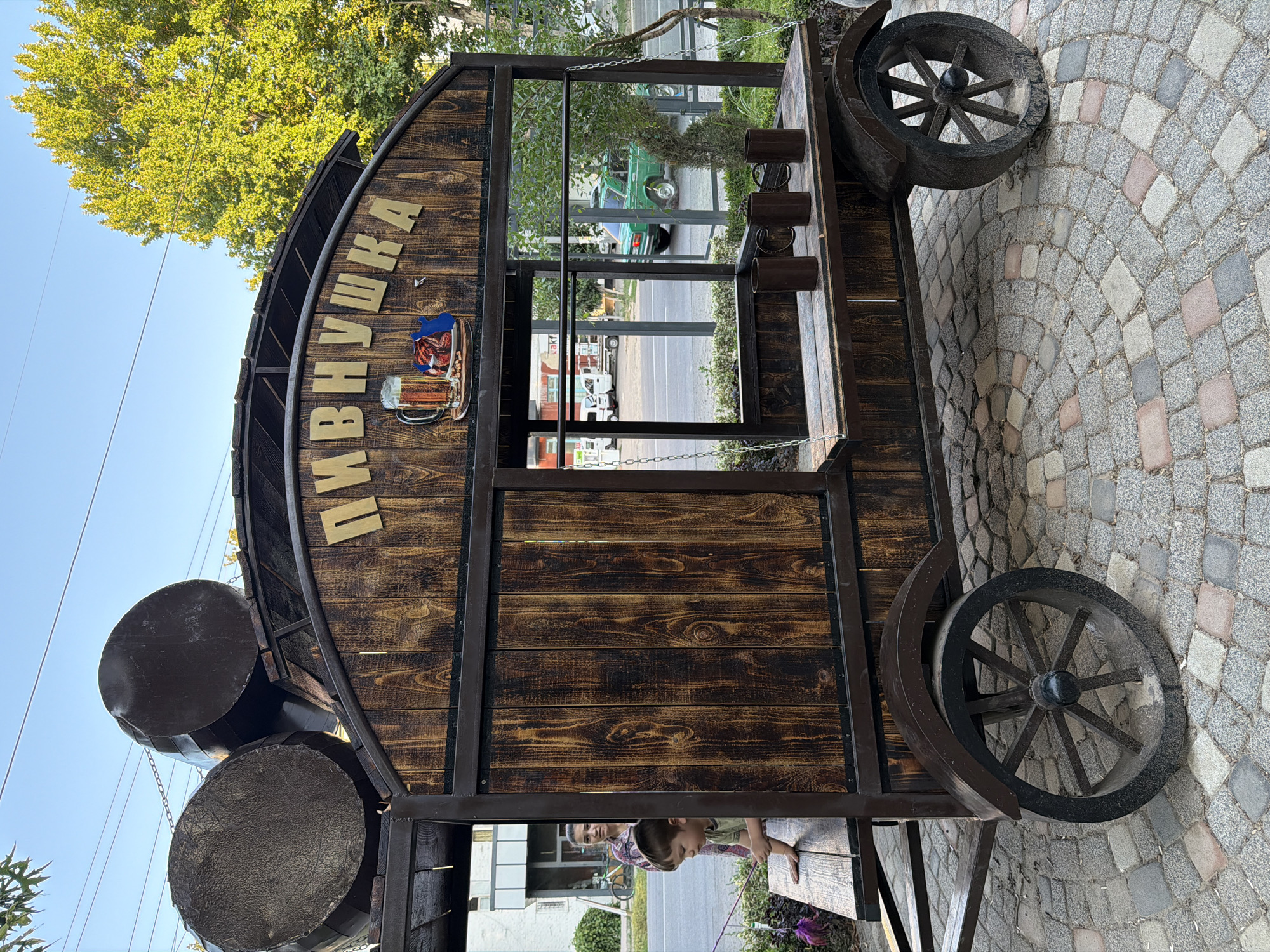 A wooden cart with a rustic design, resembling a beer stall or kiosk, is parked on a cobblestone pavement in Chirchik, Uzbekistan. The cart is adorned with large, dark barrels on its roof and a sign that reads "ПИВНУШКА" (Pivnushka), which translates to "Beerhouse" or "Little Beerhouse" in Russian. Below the sign, there is a graphic of a beer mug and some food items.
A young boy, wearing a light green shirt, is leaning over the counter of the cart, appearing to be engaged with something outside the frame. A woman, dressed in a patterned shirt, stands behind him, partially visible. The cart features wooden planks for its walls and shelves, and large, spoked wheels that give it a vintage appearance.
In the background, across a street, there are buildings, trees, and other vehicles, including a distinctive green Soviet-era car. The sky is clear and blue, indicating fair weather. The overall scene suggests an outdoor setting, possibly a park, a street market, or a public area in the city.
