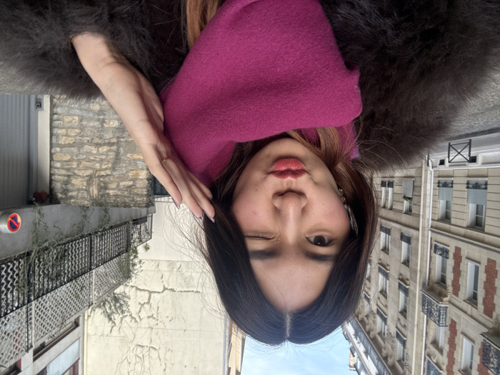 A young East Asian female is depicted in a close-up, front-facing image, winking her right eye and pursing her lips in a kiss gesture. She has long, dark hair with lighter ends, parted centrally. She wears a dark, possibly fur or faux-fur, coat and a bright fuchsia pink scarf wrapped around her neck. Her left hand is raised near her head, fingers slightly spread, revealing light-colored nail polish and a thin gold ring on her left ring finger.
The background features typical Parisian urban architecture. To the left, a multi-story Haussmannian building with light-colored stone facades, multiple windows, and black wrought-iron balconies is visible. A cobblestone street and a low black metal barrier are present at the bottom left. To the right, a stone wall base supports an elevated structure featuring a black metal railing and a white lattice screen, partially overgrown with green vines. Behind this, a plain, light-colored building wall exhibits visible cracking. A red and white circular "No Parking" sign (interdiction de stationner) is mounted on a grey surface, likely a garage door or wall, in the lower right. The scene is illuminated by natural light under a bright sky, indicating an outdoor location in Paris, France.