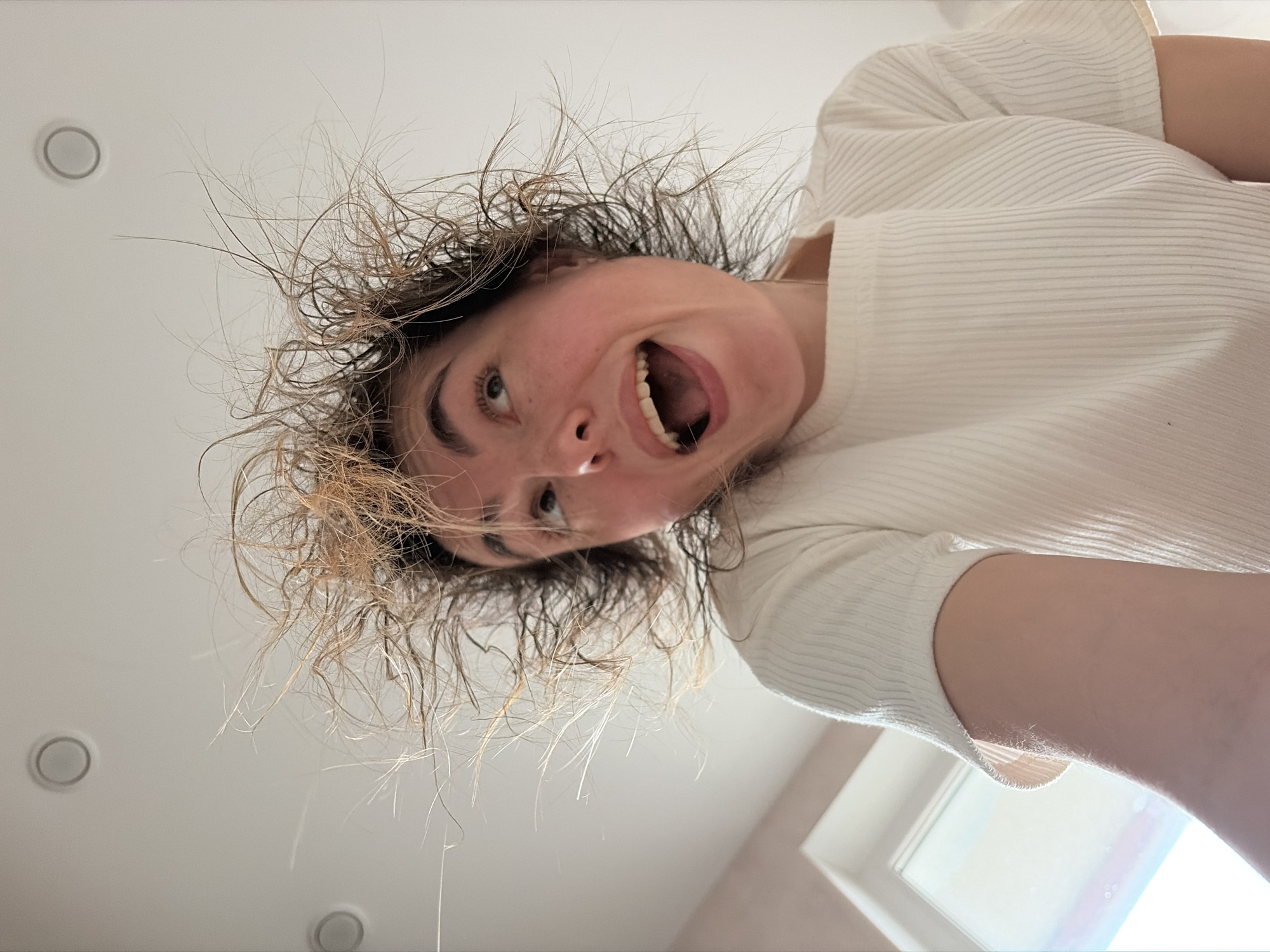 This close-up, low-angle image, taken in Izhevsk, Russia, features a young person, likely female, looking down towards the camera with a highly expressive and energetic demeanor. Their head is tilted, and their mouth is wide open, revealing both upper and lower teeth, suggesting a shout, laugh, or scream. Their eyes are open wide and appear to be looking upwards and slightly to the side.
The subject's hair is a notable feature, appearing disheveled, frizzy, and wild, with individual strands standing out in multiple directions, suggesting it might be wet or recently styled into a chaotic state. The hair color seems to be a mix of light brown or blonde with darker roots. The person is wearing a light-colored, possibly white or off-white, short-sleeved top with a ribbed texture.
The background is a plain, light-colored ceiling or wall, indicating an indoor setting. Two circular recessed lights are visible on the ceiling, one in the upper left and another in the lower left. A bright window or light source is partially visible in the lower right corner, providing natural illumination to the scene. The overall impression is one of playful exuberance or surprise. There is no visible text.
