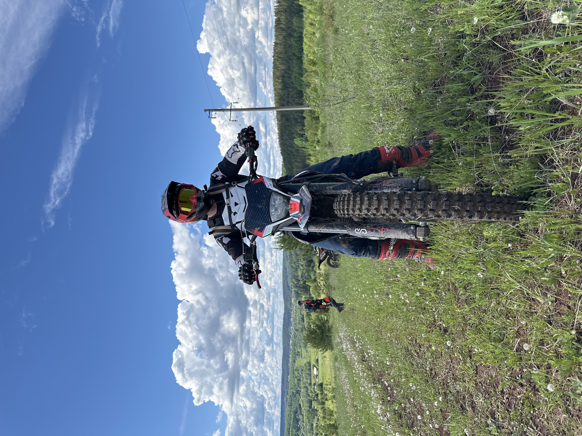 The image depicts a person on a dirt bike in a grassy field on a sunny day. The person is wearing a helmet with yellow goggles, a black and white racing jersey, and red boots. They are holding the handlebars of the dirt bike, which is positioned at a slight angle. In the background, there is a forest, a blue sky with white clouds, and power lines. Another person on a bike is visible in the mid-ground. The location appears to be near Ilyinsky, Russia, based on the landscape and vegetation. The scene appears to be an off-road riding area.