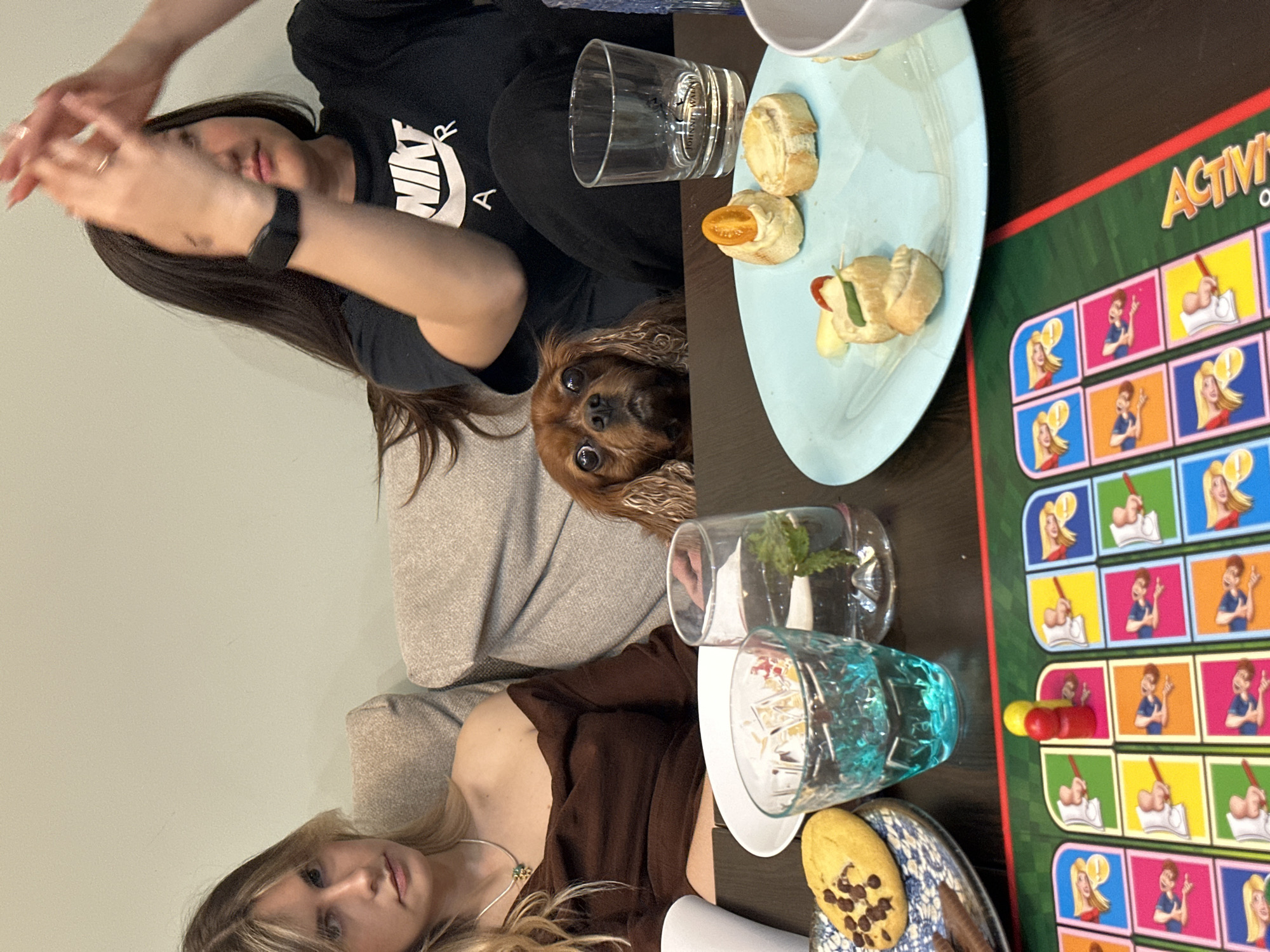 The image captures a casual indoor gathering featuring two young women and a dog, surrounded by snacks, drinks, and a board game on a dark wooden table. The scene suggests a relaxed social setting.
In the upper part of the frame, a woman with long dark hair is partially visible. She is wearing a black t-shirt with "NIKE" printed in white. Her hands are raised and slightly blurred, suggesting motion or an expressive gesture. Below her, a small, brown-furred dog, possibly a Cavalier King Charles Spaniel, peeks out from behind her, looking directly at the viewer with wide, curious eyes. The dog is positioned on a light-colored sofa or cushion.
In the lower part of the frame, another young woman with blonde hair is seen from the chest up, wearing a brown top and a silver necklace. She looks calmly towards the camera.
On the table, several items are arranged. A light blue oval plate holds small bread rolls, some topped with cherry tomatoes and others with cheese and greens, indicating appetizers or finger foods. There's a clear glass with "Johnnie Walker" branding visible, and another clear glass containing a sprig of mint or similar greenery. A textured blue glass holding a drink is also present. A blue and white patterned plate holds a chocolate chip cookie and what appear to be chocolate stick snacks. Adjacent to these items, a colorful board game is spread out, with the word "Activi" (likely "Activity") clearly visible on its green border. The game board shows various cartoon illustrations of people engaged in different actions.
The overall atmosphere is informal, suggesting friends enjoying leisure time together with food, drinks, and a game. The lighting appears to be indoor, without strong indicators of a specific time of day beyond a casual hangout.
