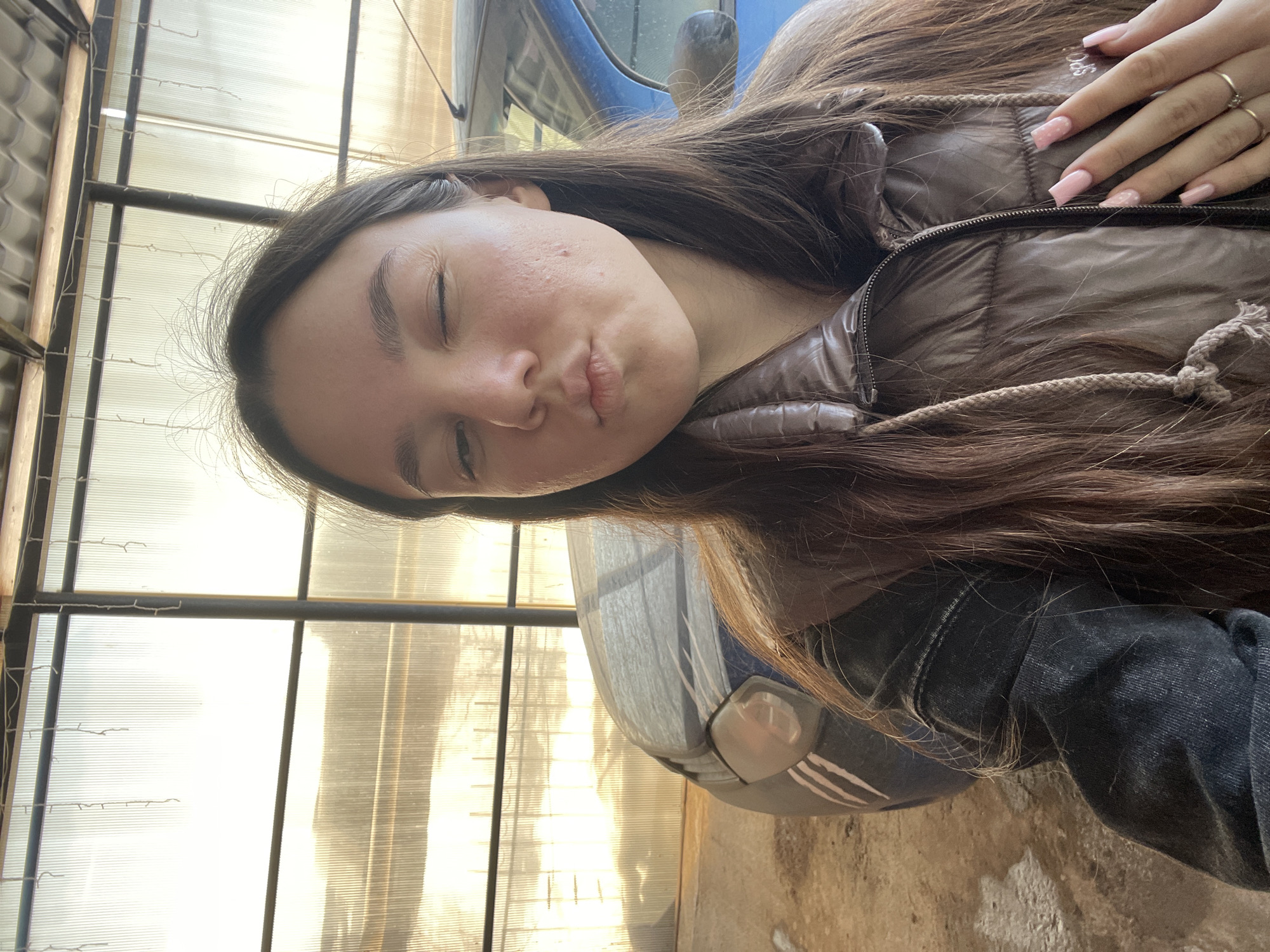 The image depicts a young female subject captured in a self-portrait within a semi-enclosed structure. The location is stated to be in the city of Фоки, Russia; however, no identifying markers within the image confirm this specific geographical detail.
**Subject:** A female, estimated to be a young adult, with long, dark brown hair parted centrally. She exhibits a wink with her right eye and her lips are puckered. Visible on her left cheek are several small blemishes. She is dressed in a shiny, brown, quilted puffer vest worn over a dark-colored garment, possibly a denim jacket. Her left hand is raised to chest level, displaying light pink, square-shaped manicured nails with white speckles, and a thin gold-colored ring on her left ring finger. A partial white logo, possibly "SPO," is visible on the front-right side of her vest.
**Environment and Objects:** The background consists of a bright, translucent corrugated panel wall, likely constructed from plastic or polycarbonate, which diffuses sunlight. This wall is supported by dark metal structural elements, including vertical posts and horizontal beams. Above the translucent wall, a portion of a grey corrugated metal roof is visible. Several thin, white, string-like objects hang vertically from an overhead beam. To the subject's left, the front-left portion of a blue automobile is visible, appearing to be a compact car or hatchback, with its headlight and part of the front bumper discernible. The ground in the foreground is an uneven surface, possibly concrete or dirt, with light-colored patches. The overall lighting suggests an outdoor or brightly lit covered area.