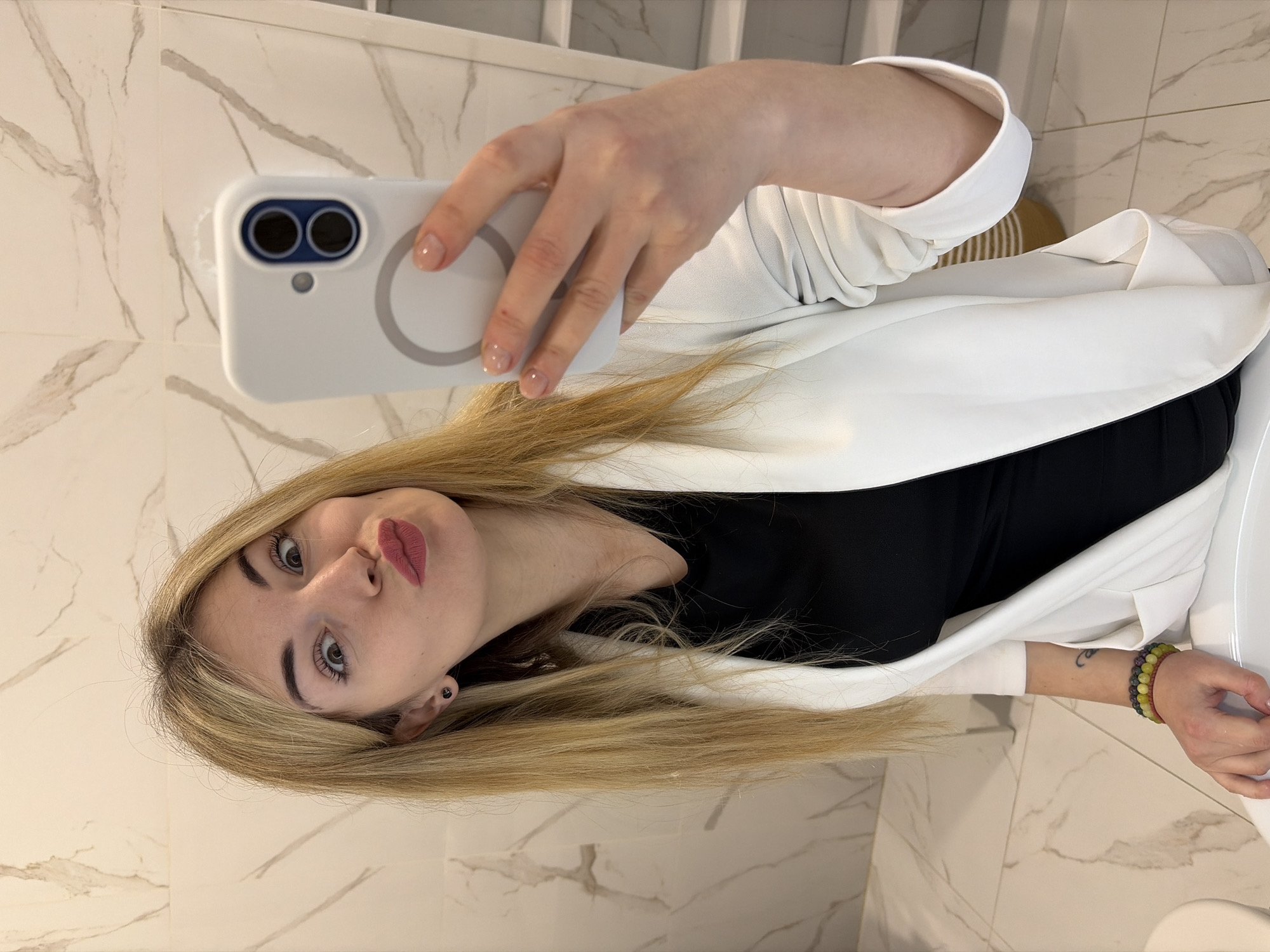 A woman is taking a selfie in a bathroom. She is wearing a white blazer over a black top. Her hair is blond. She is holding a white smartphone. The background is a white marble wall. Location in Budva, Montenegro, cannot be determined from the image.