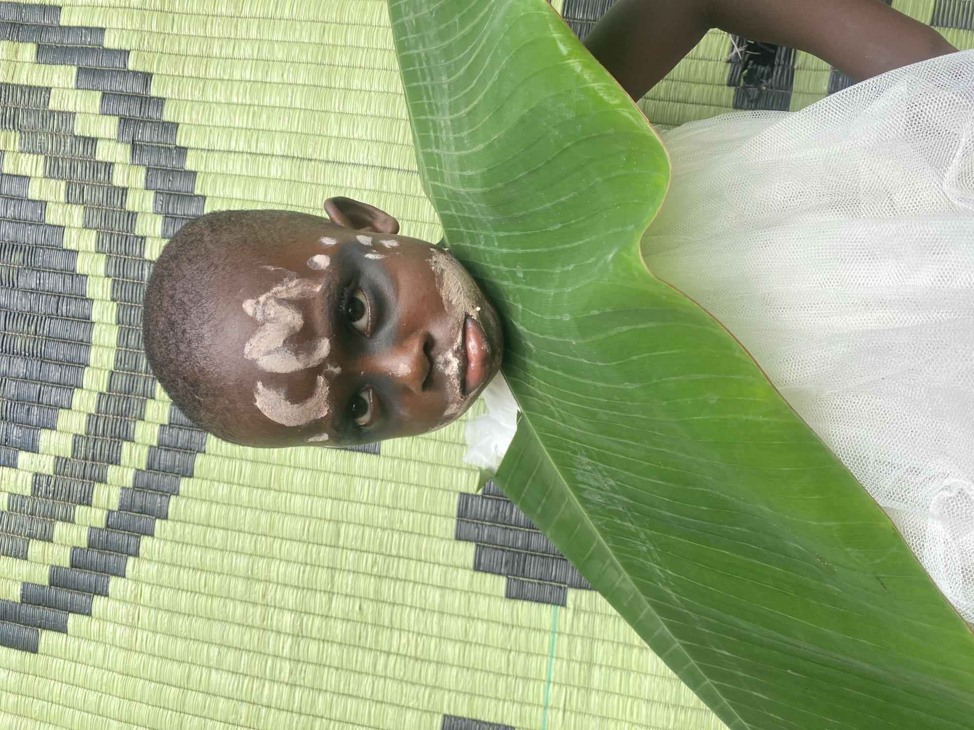A child with face paint is lying on a mat with a banana leaf in Ufu Esioni, Nigeria. The mat is green and gray with a repeating geometric pattern. The child has dark skin and white face paint in circular patterns. They are wearing a white, sheer garment. A large, bright green banana leaf is positioned near their head and upper body. The child is lying on their back.