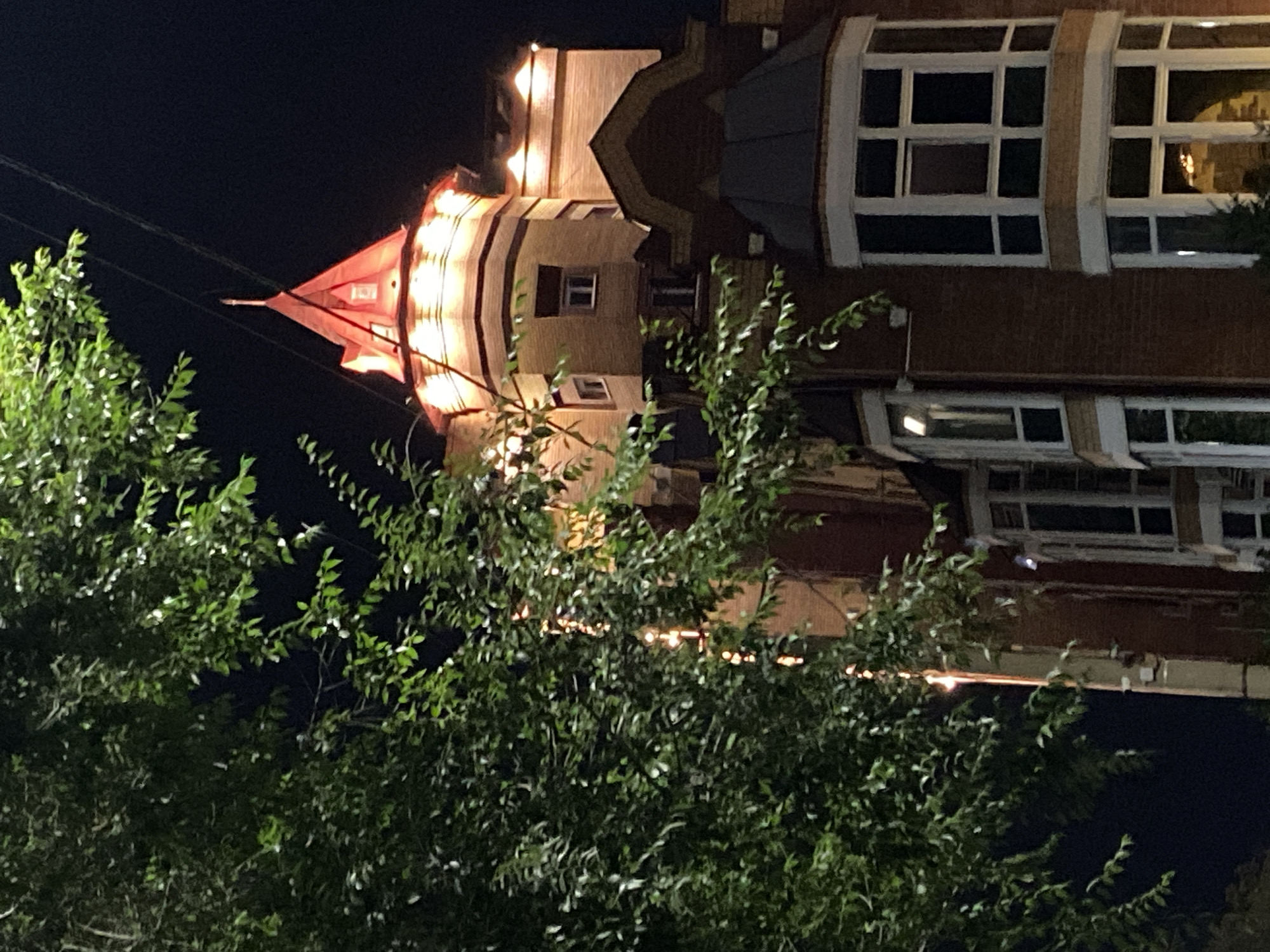 A close-up, low-angle, night-time, outdoor shot captures the upper section of an ornate, red-brick building with a pointed, illuminated red roof, resembling a tower. The building features multiple windows, some appearing as bay windows with multiple panes. A large, leafy green tree with scattered bright lights along its branches partially obscures the left side of the building. The background is a dark, clear night sky. The image was taken on October 27, 2022, in Blagoveshchensk, Russia.