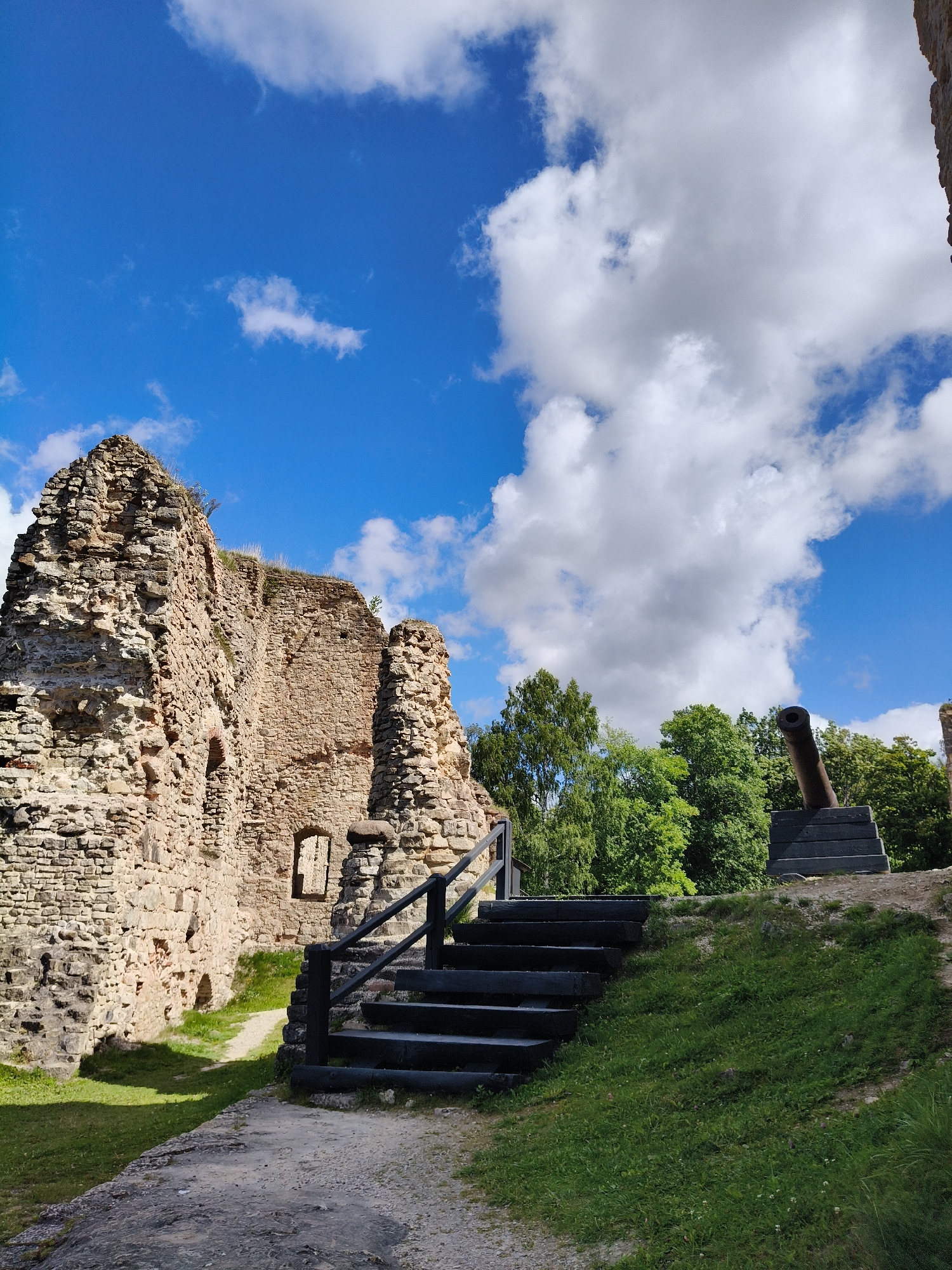 The image shows the ruins of Koknese Castle in Koknese, Latvia, under a bright blue sky with fluffy white clouds. In the foreground, a stone path leads towards a set of dark wooden stairs with a railing, ascending a grassy slope. To the left, the crumbling stone walls of the castle dominate the scene, revealing ancient architecture with arched openings. Further up the slope, a weathered, dark-colored cannon is mounted on a wooden base, positioned overlooking the landscape. Lush green trees form a dense backdrop behind the ruins and the cannon. The overall impression is one of historical preservation and a serene, sunlit day.
