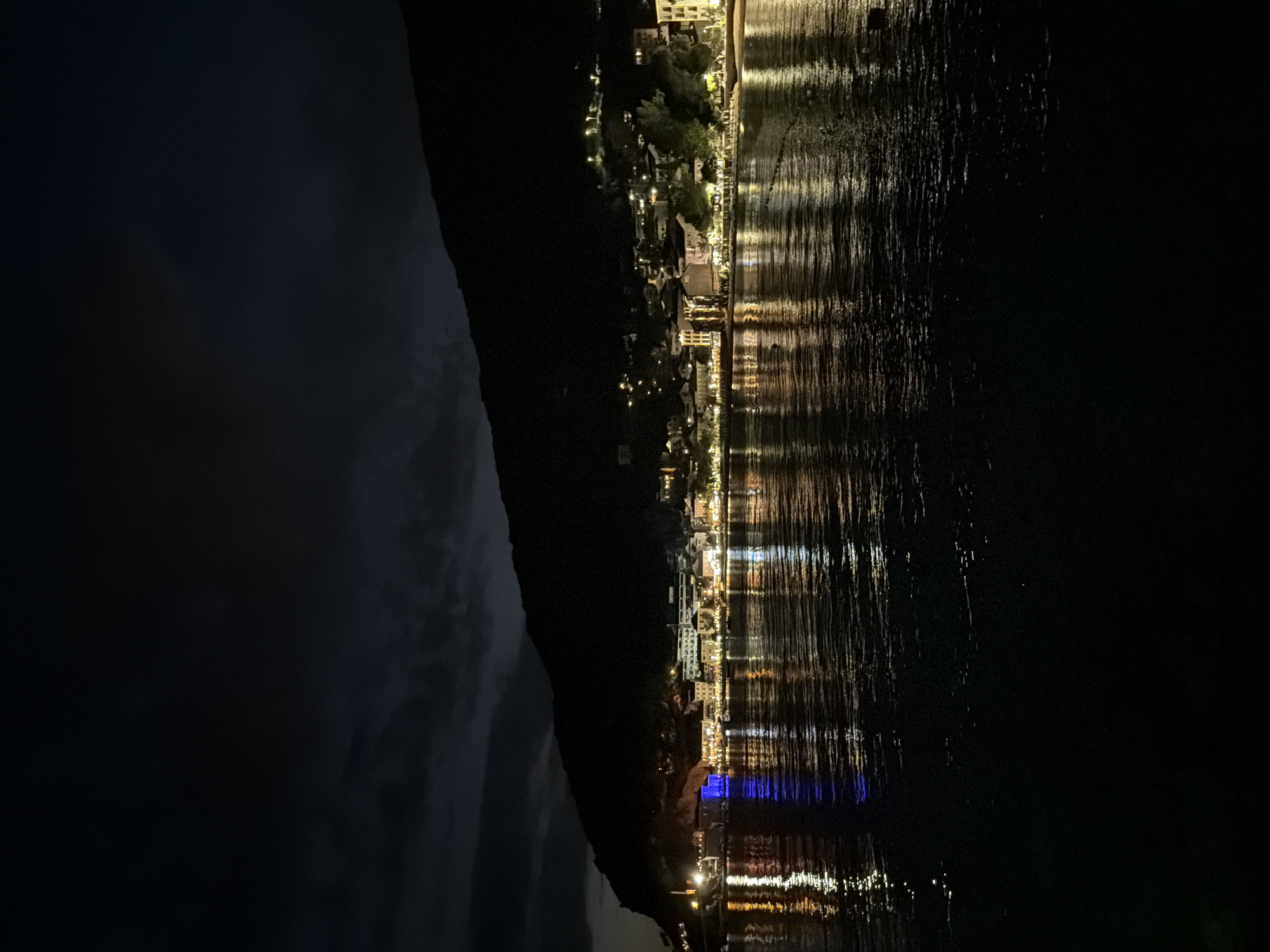 The image depicts Petrovac centar, Montenegro, at night, from the perspective of the sea. The dark sky is partially obscured by faint cloud formations, visible above a prominent, unlit mountain that forms a dark silhouette spanning the upper half of the background. Along the base of the mountain, a densely built coastal town is illuminated by numerous artificial lights. These lights, originating from buildings, streetlights, and possibly commercial establishments, cast bright reflections across the dark, rippled surface of the water in the foreground. A distinct, intense blue light emanates from a structure on the left side of the town, creating a prominent blue reflection on the water. Other lights appear yellow or white, contributing to a tapestry of broken light streaks on the water's surface. No individuals or specific activities are discernible. The overall scene conveys a nocturnal urban coastal landscape with significant light pollution reflecting on the water.