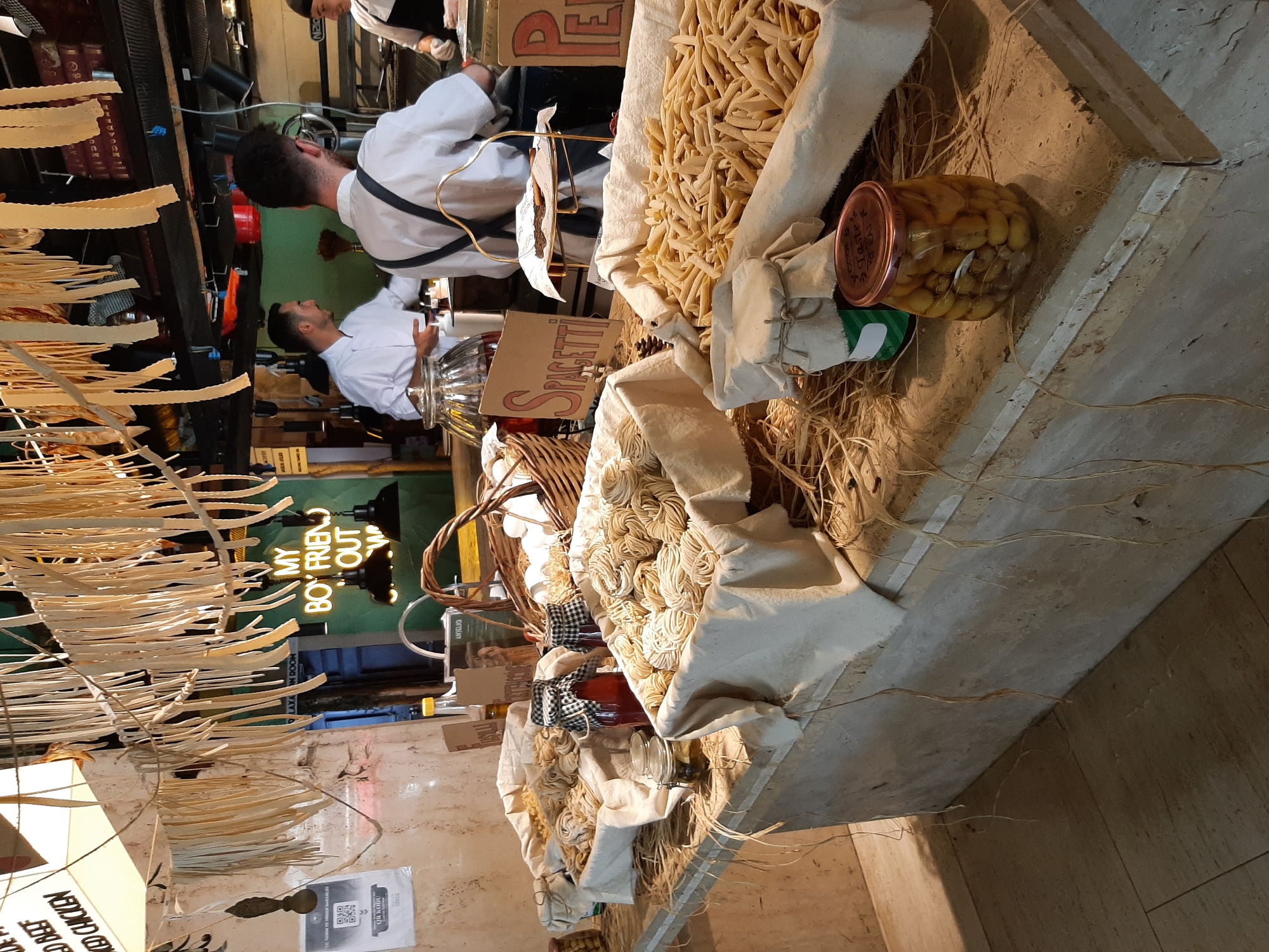 This image captures a vibrant food stall, likely a pasta shop, in Alanya, Turkey. The stall features a rustic aesthetic with hanging pasta strands and straw detailing. In the foreground, several large baskets overflow with various types of pasta, including spaghetti and penne. Jars of preserved food items, such as olives, add to the display. Behind the counter, three individuals, presumably staff, are engaged in food preparation. Two men in white chef coats are visible, one interacting with a customer or colleague, and another with his back to the camera. A third person, also in a chef's uniform, is partially visible to the right. The environment suggests a busy, open-air market or restaurant setting, with a neon sign in the background reading "MY BOYFRIEND OUT".