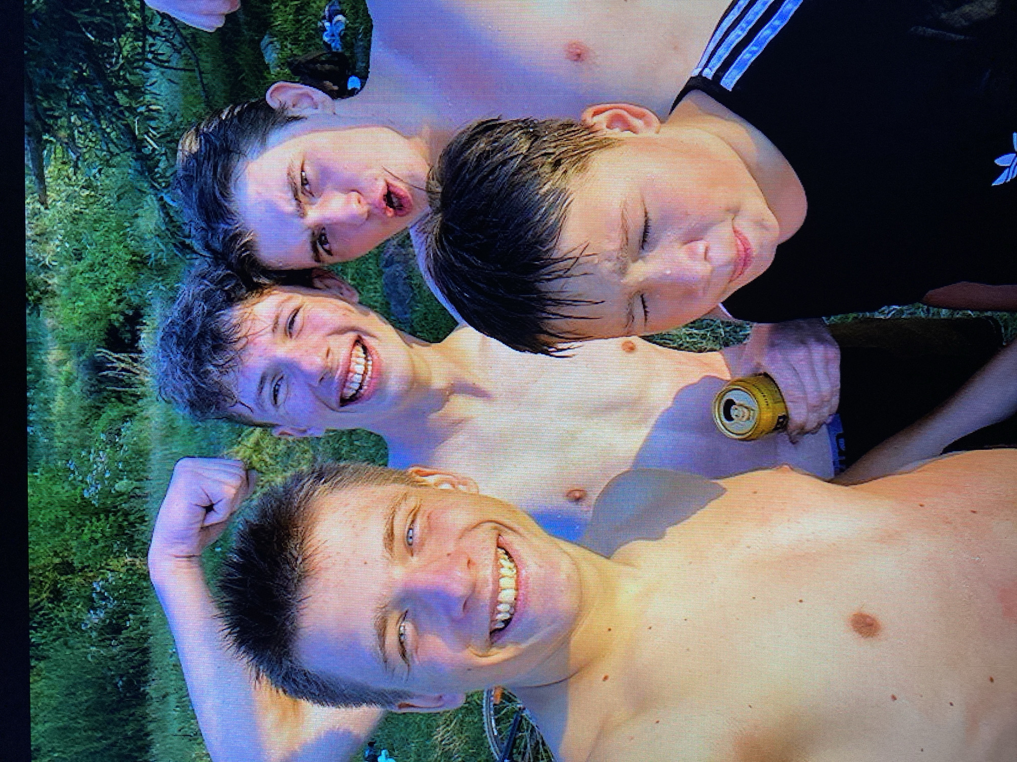 The image shows a group of four young males, appearing to be teenagers, posing for a selfie outdoors. The background consists of dense green foliage, suggesting a natural or park-like setting. The individuals are shirtless, with one holding a can, possibly a beverage. Their expressions range from smiling to a more serious pout. The lighting suggests it is daytime, likely with natural sunlight. Without further information or visual cues, the specific location within the city of Poznan, Poland, cannot be determined from the image alone.