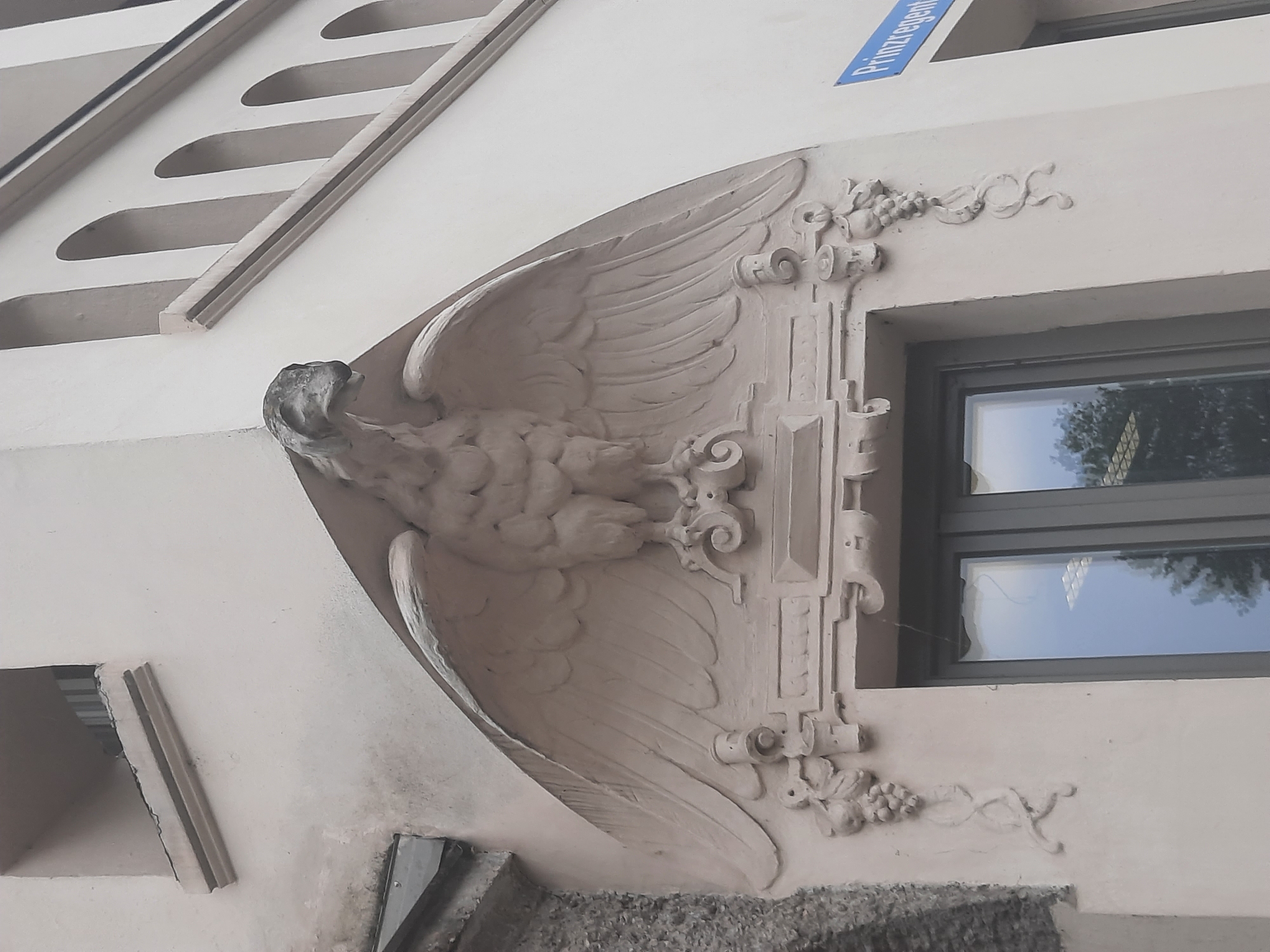 A building facade in Rosenheim, Germany, features a relief sculpture of an eagle with spread wings above a window. The eagle sculpture is made of stone and is adorned with ornate carvings on either side, including grapevines and fruit. To the right of the building, a blue street sign reads "Prinzregenten". The building's architecture includes arched windows and decorative moldings. The overall scene depicts architectural ornamentation on a building's exterior.