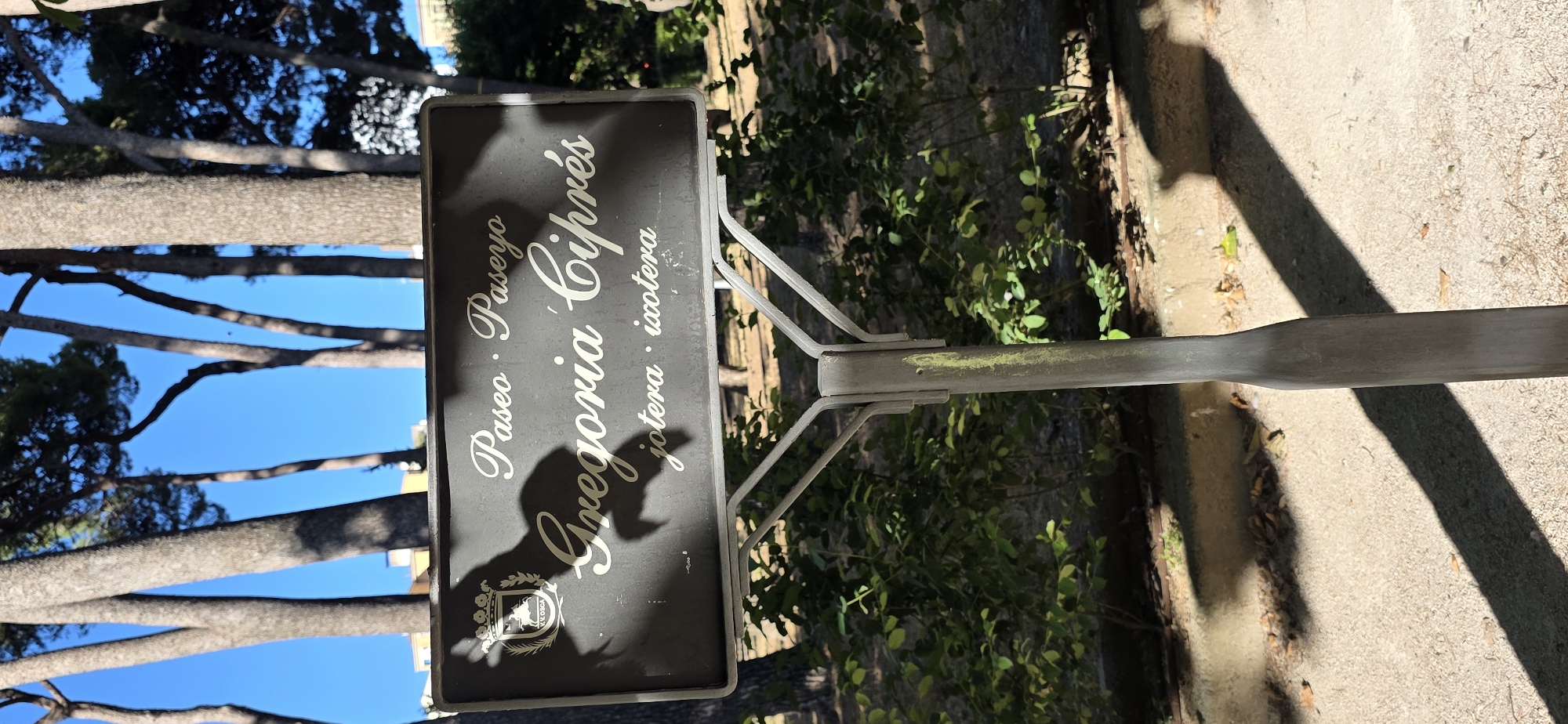 The image shows a dark sign with white text, mounted on a metal pole. The sign has a crest at the bottom left. The text appears to be in Spanish and reads: "Paseo · Paseyo Gregoria Ciprés · ixotera". The sign is positioned outdoors, with trees and a bright blue sky visible in the background. The lighting suggests it is daytime, likely sunny. The ground appears to be paved. The overall setting seems to be a park or a street with trees. The sign's inscription indicates it's a place marker, possibly for a promenade or street named "Gregoria Ciprés". The presence of a crest suggests it might be a historical or officially designated area. The orientation of the image is portrait, and the sign is angled towards the viewer.