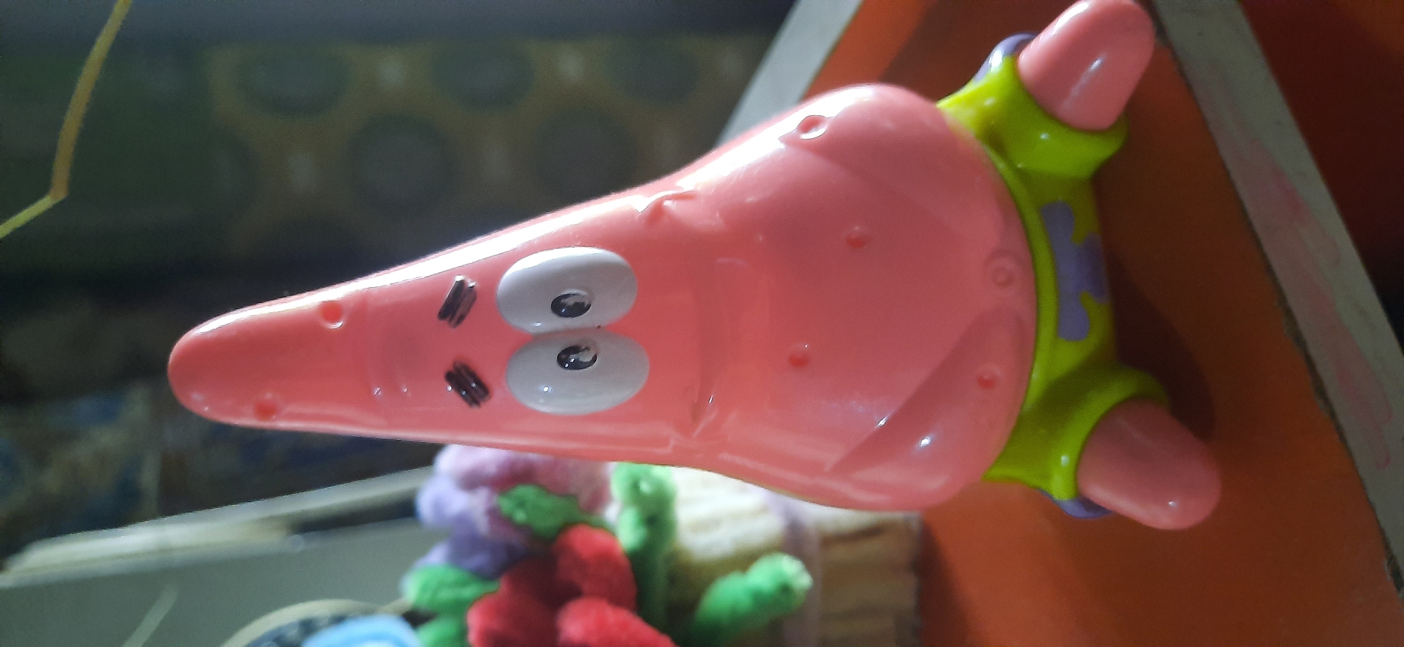 A close-up, eye-level, vertical shot features a pink plastic toy resembling the character Patrick Star from SpongeBob SquarePants, positioned centrally on an orange surface. The toy, with its distinctive star shape, has two large white eyes with black pupils and thick black eyebrows. It is wearing green shorts with a purple design.

To the left and slightly behind the toy, a blurred arrangement of plush toys is visible. These appear to be colorful, with hints of red, green, and purple. The background is out of focus, displaying a patterned wallpaper or fabric with dark green and brown tones, suggesting an indoor setting.

The lighting is moderate, with some reflections visible on the plastic toy, particularly on its head and eyes. The overall impression is that of a toy displayed on a table or shelf. The origin of the image is identified as Cisauk, Indonesia, suggesting the photograph was taken in this location. No people are present in the image.