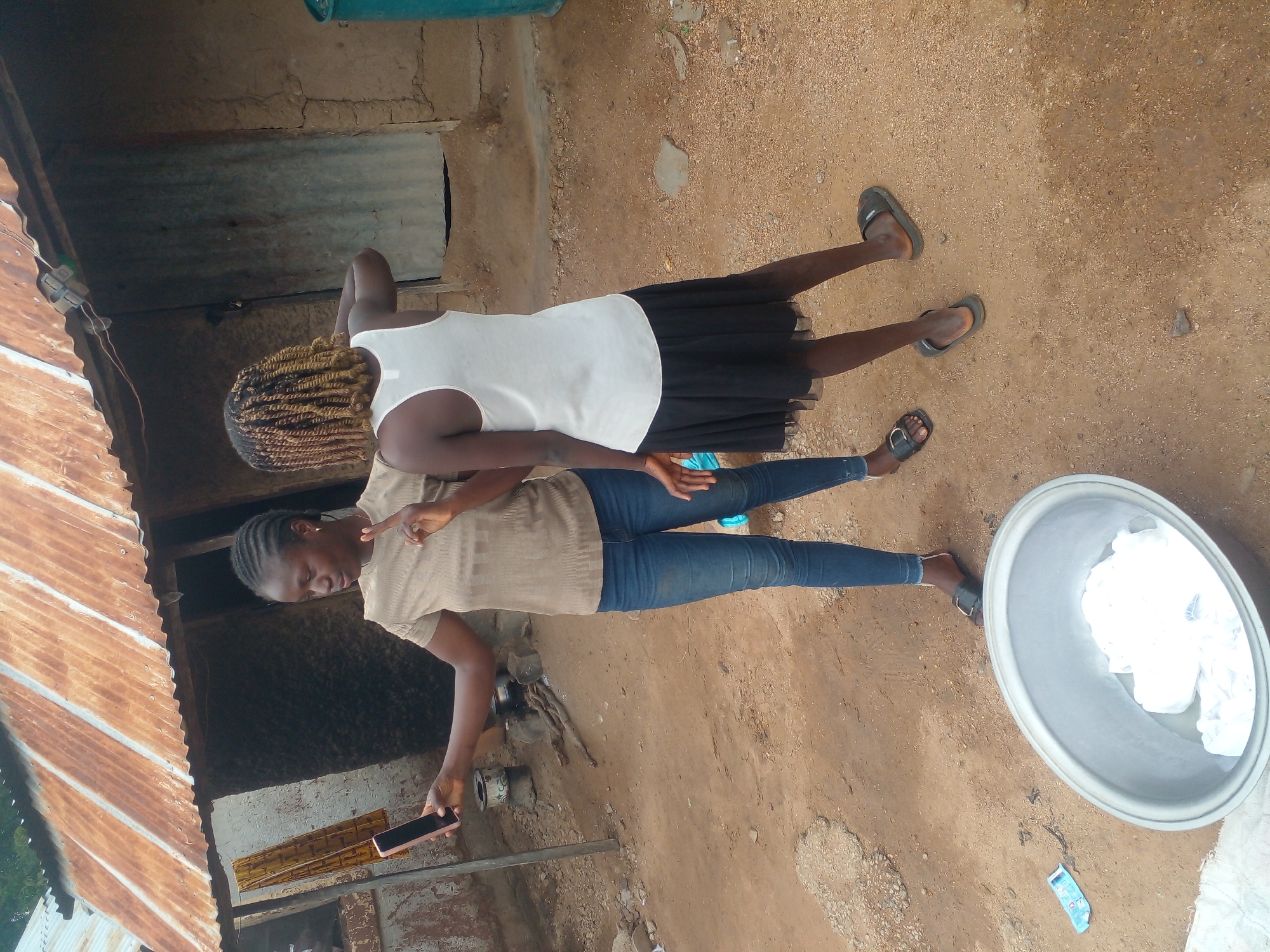 Two young women are standing outdoors, possibly in a village setting. The woman on the left is holding a pink smartphone in her left hand, taking a selfie, and has her right hand raised with two fingers extended in a peace sign. She is wearing a light brown, textured short-sleeved shirt and blue jeans. The woman on the right is facing away from the camera, wearing a white tank top and a black skirt. The background features a rustic building with a corrugated metal roof that appears to be weathered and rusted. The ground is dirt and sand. In the foreground, a large, grey basin is filled with water and white cloth, suggesting laundry is being done. There is also a small blue packet on the ground near the basin. The overall scene depicts a casual moment captured in a rural or semi-rural environment.