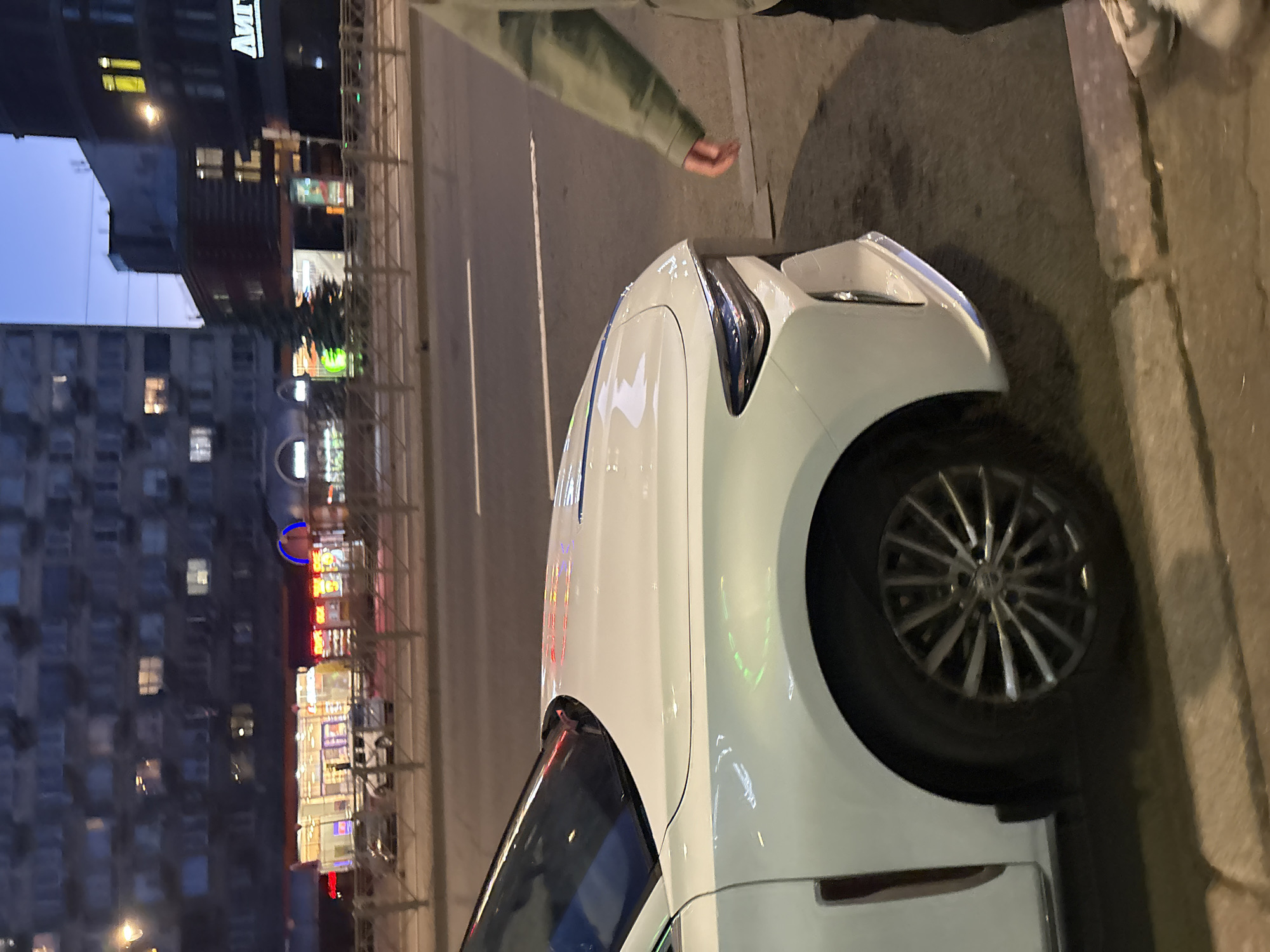 The image depicts a street scene in Saint Petersburg, Russia, at dusk or night. A white car is parked on the side of a road, facing left. The car's front fender, wheel, and part of the hood are visible. A person, wearing a light-colored jacket and dark pants, stands to the right of the car, with their right arm extended towards the vehicle. In the background, there are multi-story buildings with illuminated windows, streetlights, and a fence separating the road from the sidewalk. Signs with Russian text are visible on some buildings. The overall impression is an urban environment with some commercial activity.