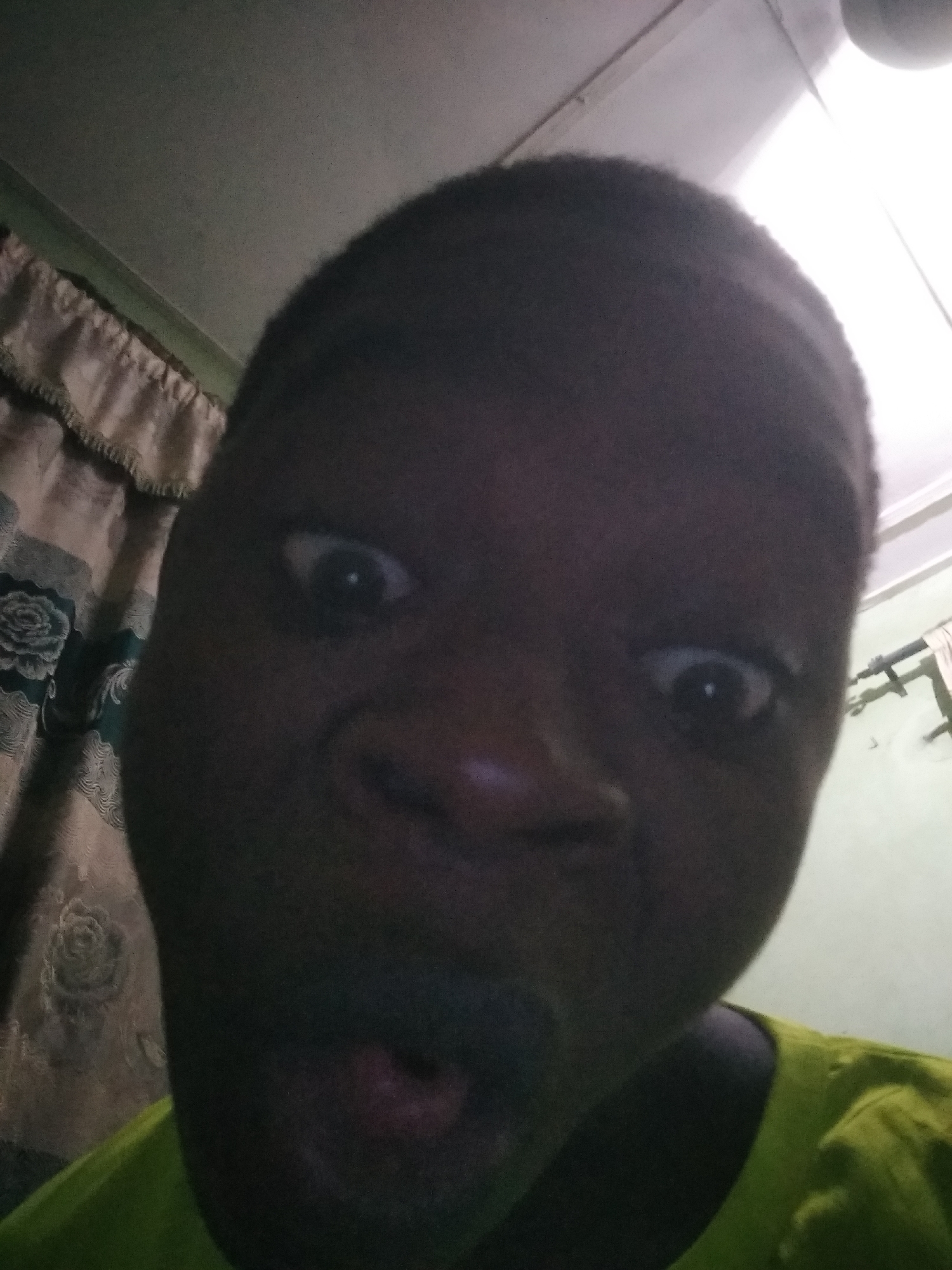 A close-up selfie taken indoors shows a dark-skinned person with wide eyes and an open mouth, expressing surprise or shock. The person is wearing a green shirt. In the background, there is a curtain with a floral pattern and a white wall. The environment suggests a domestic setting. There is no other information provided about location, activities, or interactions.