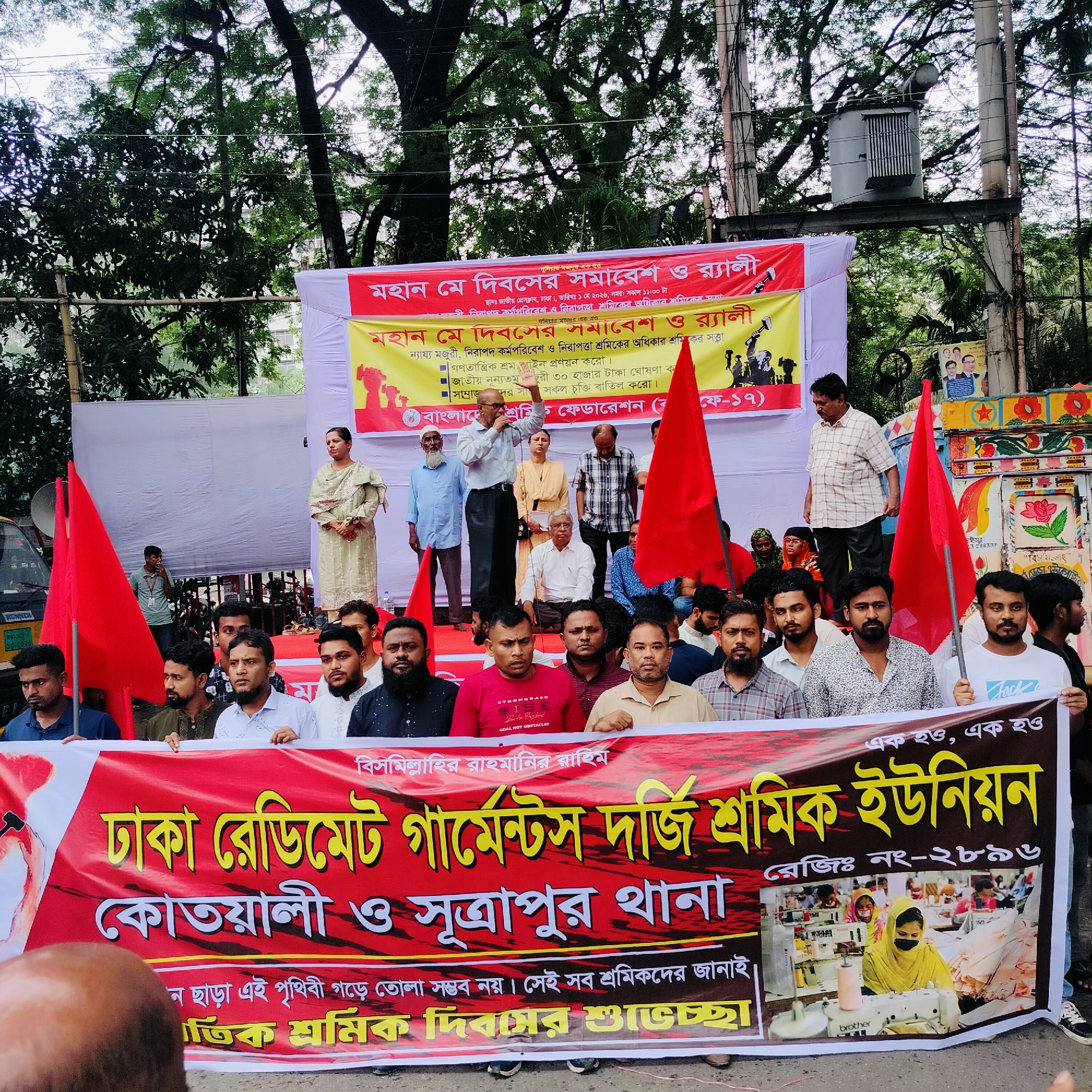 A daytime outdoor scene shows a rally of garment workers in an urban location in Unknown, Unknown. A crowd of men and a few women are gathered, some standing on a raised platform behind a large banner. The banner reads "Dhaka Readymade Garments Tailors Workers Union", in Bengali, with an image of women working at sewing machines. Above the platform is another banner also written in Bengali proclaiming "Great May Day rally" with demands for fair wages and worker rights. The speakers are using microphones, some holding red flags. The backdrop is composed of white sheeting, trees, and an electrical transformer. The crowd is holding the banner and watching the speakers. Other signs and decorations are visible, including political posters. Trees surround the area.