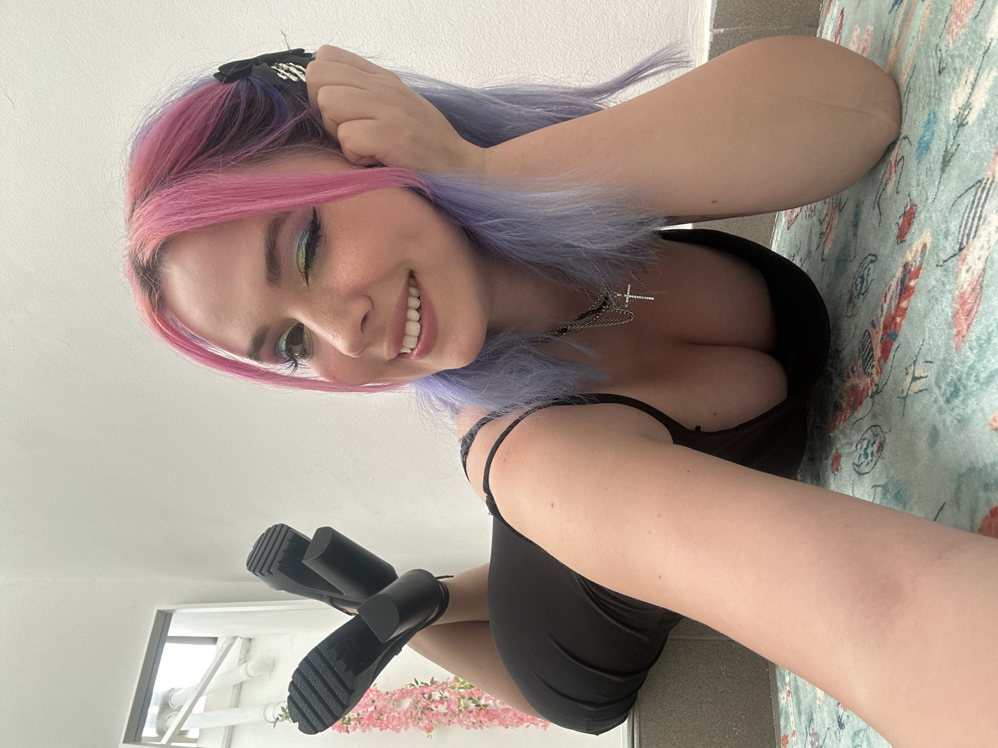 The image shows a young woman with dyed pink and blue hair, lying on a patterned rug. She is wearing a black dress and black heeled shoes. She is smiling and looking at the camera, with one hand touching her hair. A window with a view of pink flowers is visible in the background. It is impossible to determine the specific location within Zagreb from this image.