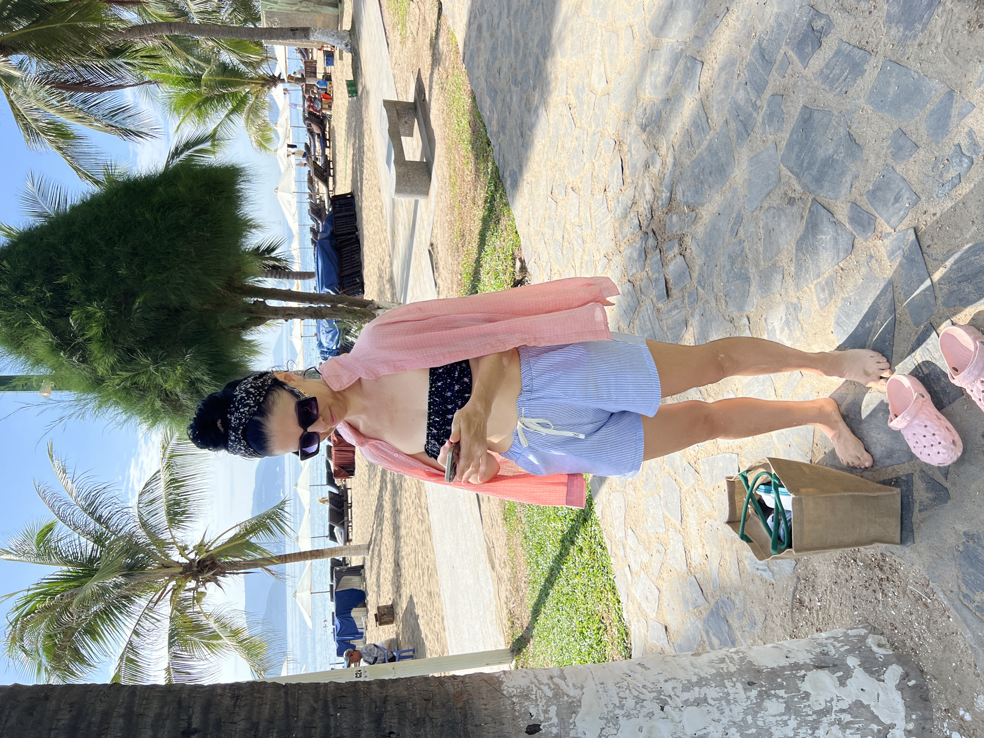 A woman is standing on a paved walkway at a beach, looking at her phone. She is wearing a black bikini top, blue and white striped shorts, and a pink open shirt. A brown canvas bag and pink clogs are on the ground near her. In the background, there is a beach with palm trees, lounge chairs, and umbrellas. The scene appears to be in Nha Trang, Vietnam.