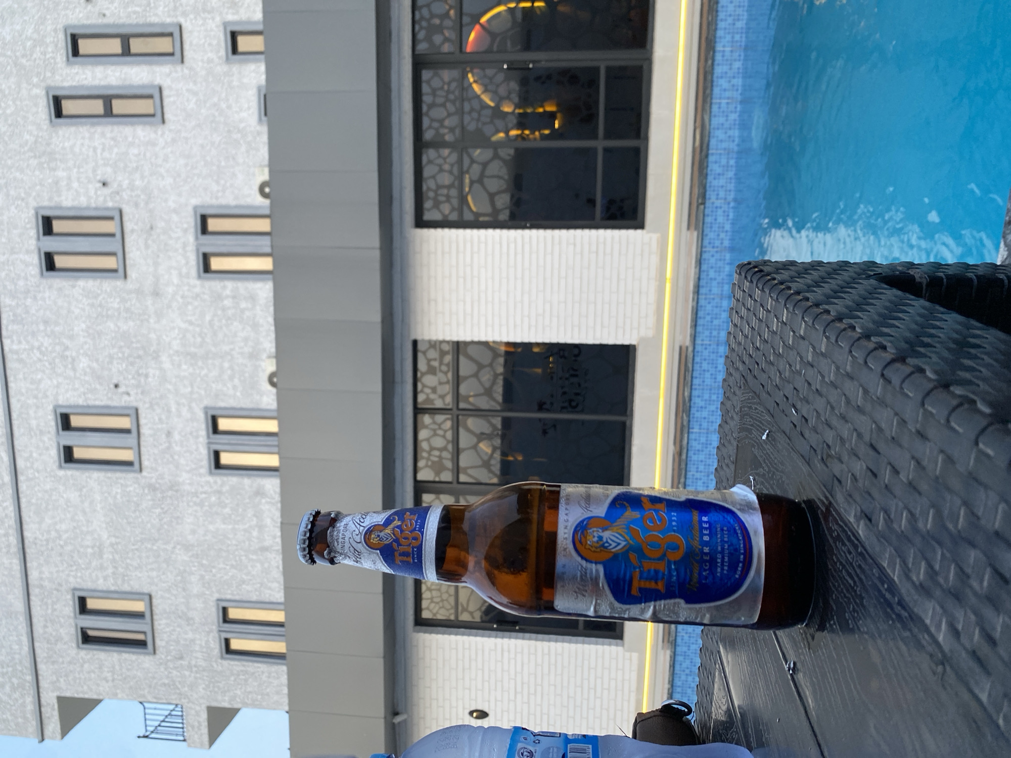 The image, taken in Kubwa, Nigeria, captures a tranquil poolside scene during what appears to be late afternoon or early evening. In the immediate foreground, a bottle of Tiger beer rests on a dark, woven outdoor table or chair. The beer bottle's label is prominent, featuring the brand name "Tiger," along with text such as "LAGER BEER," "AWARD WINNING," "PREMIUM BEER," and "SINGAPORE SINCE 1932," indicating its origin and quality. A portion of a plastic water bottle is also visible on the table.
Beyond the table, a clear blue swimming pool with tiled edges reflects the bright sky and surrounding structures, suggesting calm water. The background is dominated by a modern, multi-story building. Its lower section features a white, brick-textured facade with large dark windows that have intricate, possibly illuminated, patterns inside. The upper floors of the building are light grey, punctuated by numerous small, rectangular windows that emit a warm, golden glow from within. The overall setting suggests a leisurely environment, possibly a hotel, resort, or an upscale residential complex. The sky is bright and clear, contributing to the relaxed ambiance of the scene.