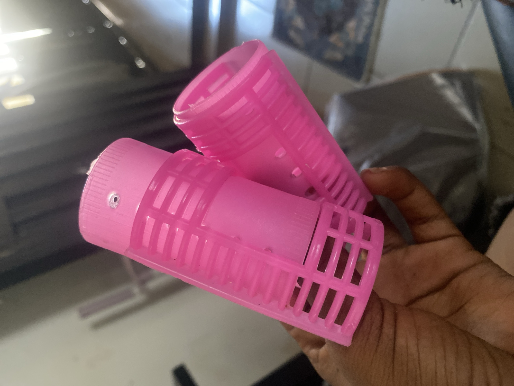 In Kubwa, Nigeria, a hand holds two bright pink plastic hair rollers. The rollers are cylindrical with a slotted design. One roller is positioned vertically, while the other is angled in front of it, partially obscuring it. The background is blurred, showing hints of indoor lighting and possibly furniture. The image is taken from a close-up perspective, focusing on the hair rollers and the hand.
