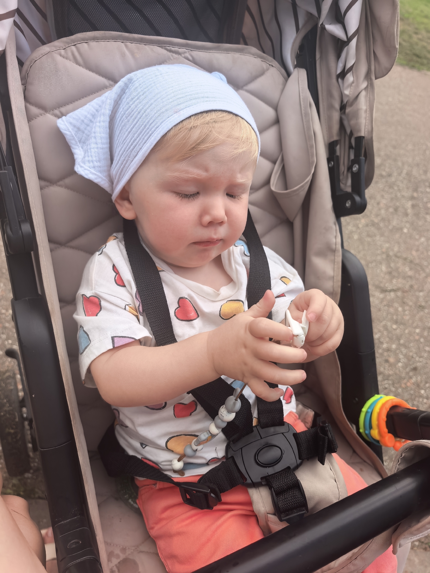 A toddler wearing a light blue headscarf and a white t-shirt with colorful heart prints is seated in a stroller. The child is holding a small white object in their hands and appears to be looking down at it. The stroller has a beige padded seat and a black safety harness. A colorful ring toy is attached to the stroller handle. The background is slightly blurred, showing a paved path and some greenery, suggesting an outdoor setting, possibly a park or a sidewalk in Sterlitamak, Russia.