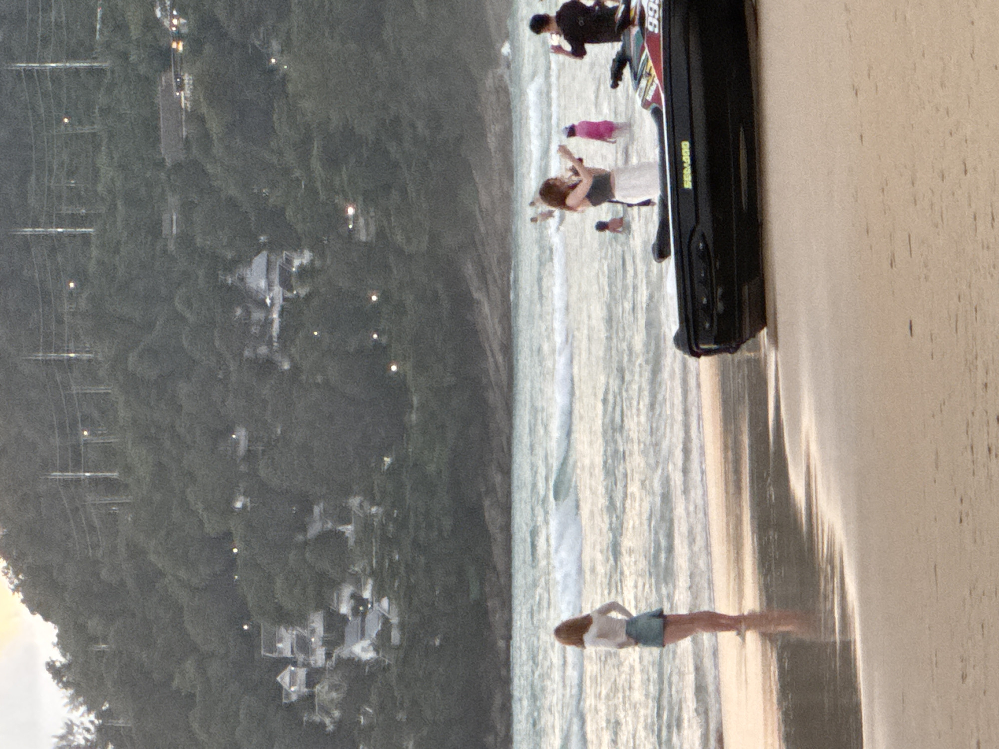 In Karon, Thailand, a beachfront scene unfolds with a view of a densely wooded hill in the background, dotted with lights from buildings and power lines. The foreground features a sandy beach with the edge of a black jet ski, identified by "SEA-DOO" branding and the number "999", resting on the sand. In the shallow water, several individuals are visible. To the left, a woman stands facing away from the camera, wearing a white long-sleeved shirt and a denim skirt. Further into the water, a woman in a dark top and a long white skirt is holding a phone, and a person in a pink garment is also present. To the right, a man in a dark shirt is seen holding up a phone. Waves are breaking in the turquoise water.